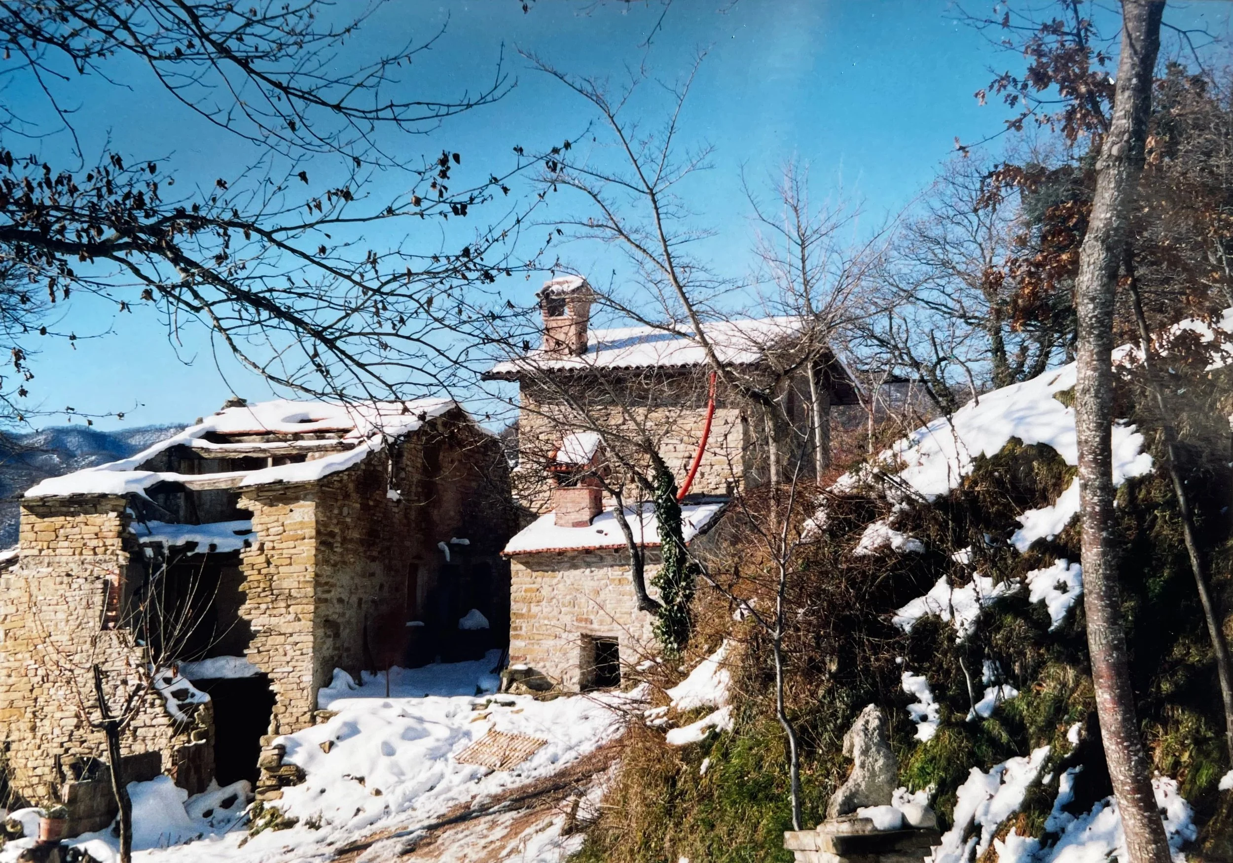 Snow-covered rustic stone house on a hillside surrounded by leafless trees under a clear blue sky.