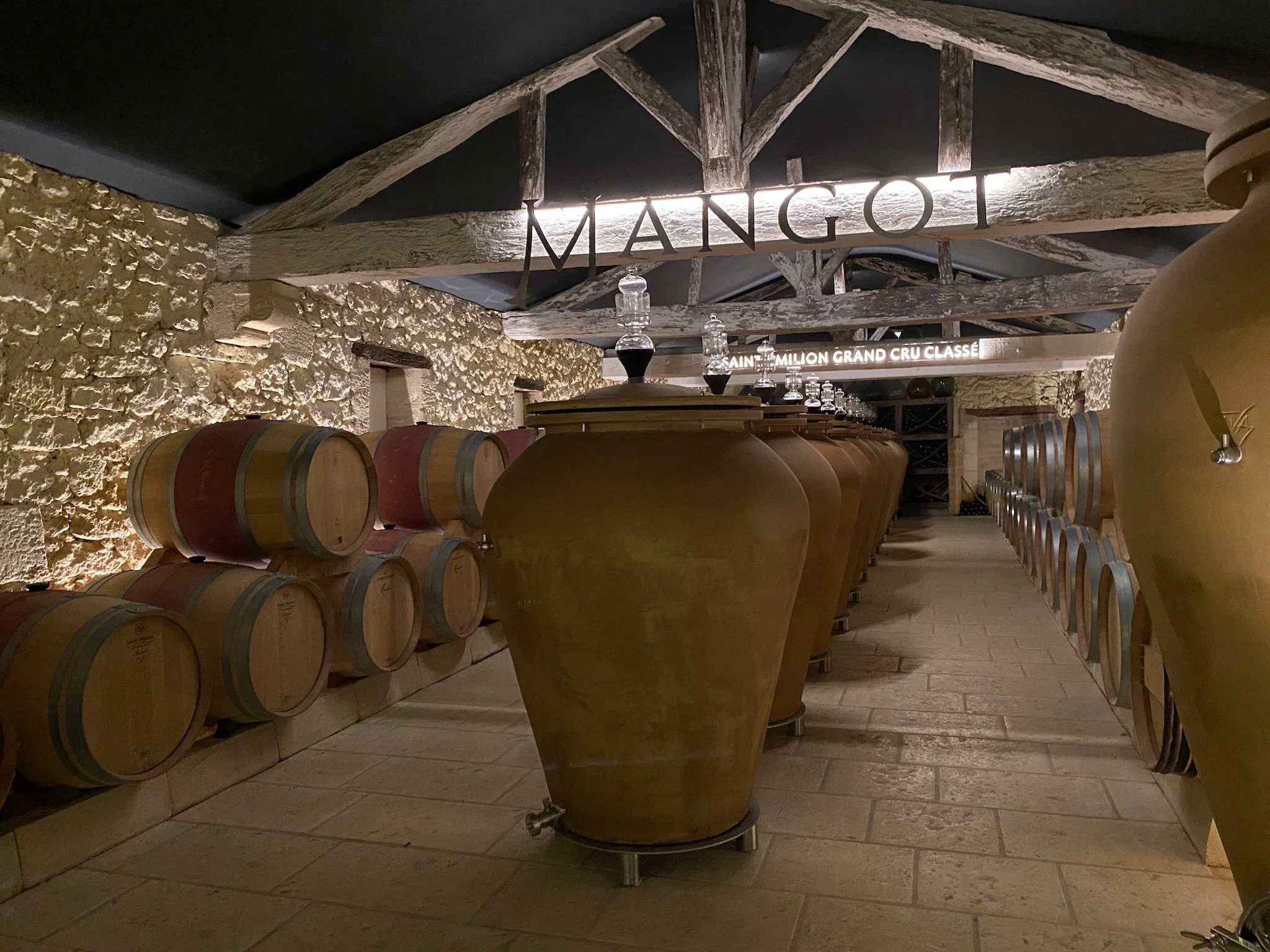 Wine cellar with barrels stacked on the left and large clay fermentation vessels on the right, illuminated signs reading "MANGOT" and "SAINT EMILION GRAND CRU CLASSÉ" overhead.