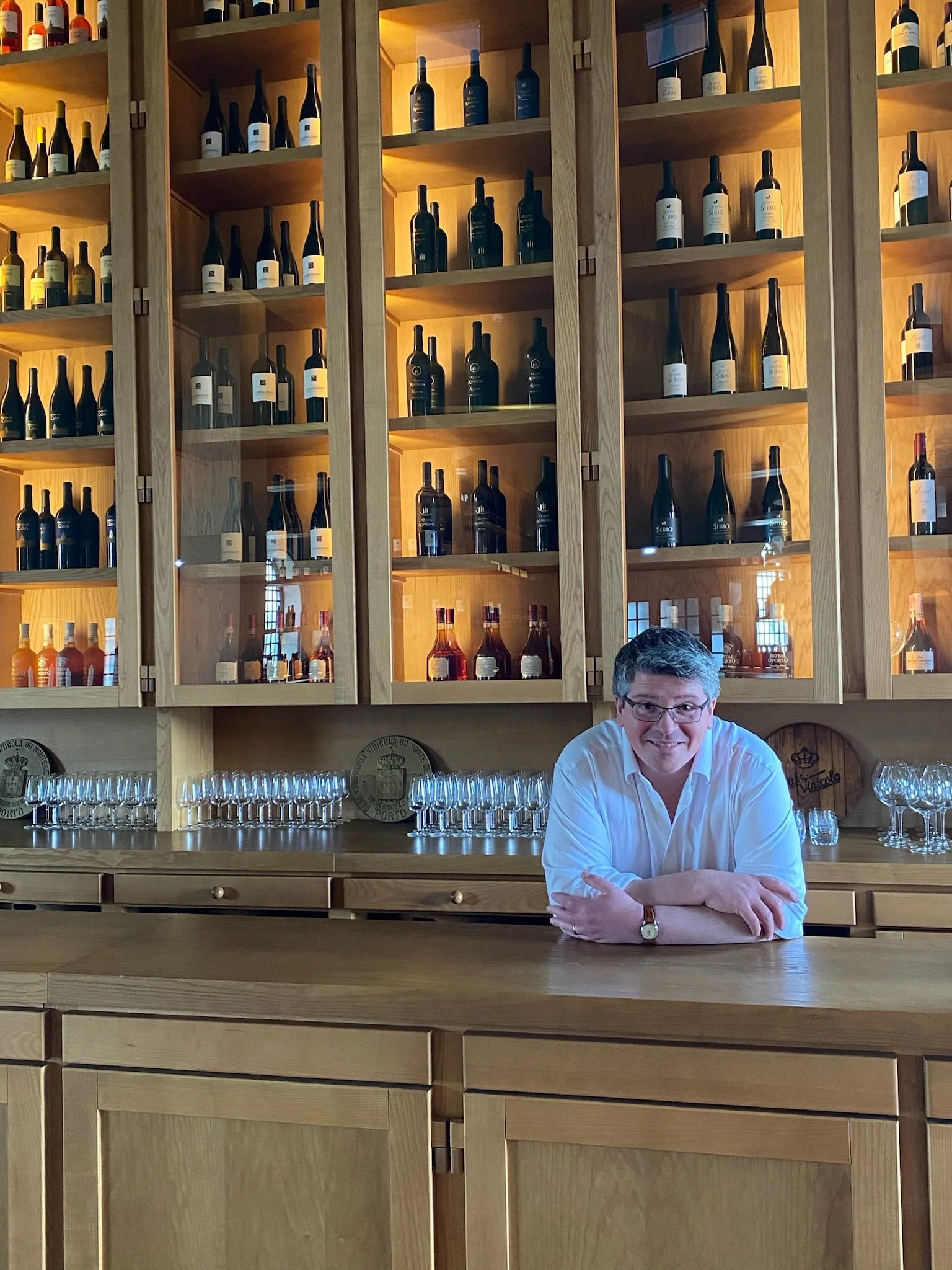 A man with glasses and a white shirt leaning on a bar counter in front of wooden shelves filled with wine bottles and glasses in a wine bar or winery