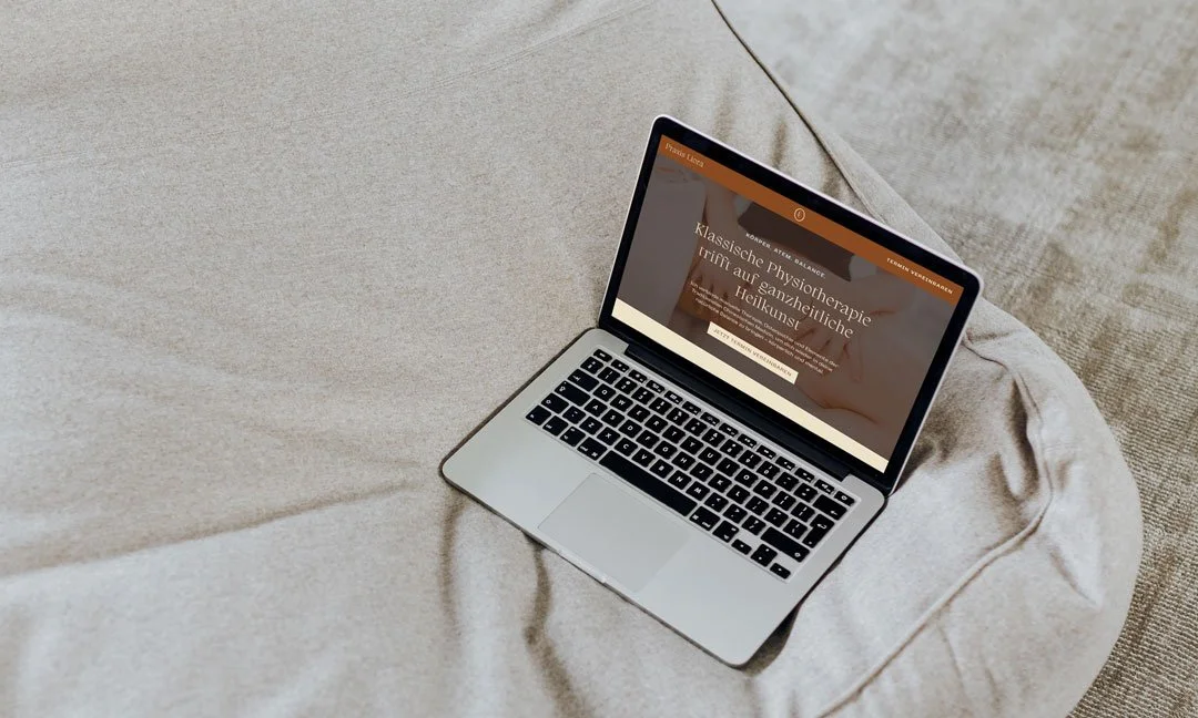 Ein Laptop liegt auf einem beige Couchkissen, zeigt eine Webseite über Physiotherapie und ganzheitliche Heilkunst.