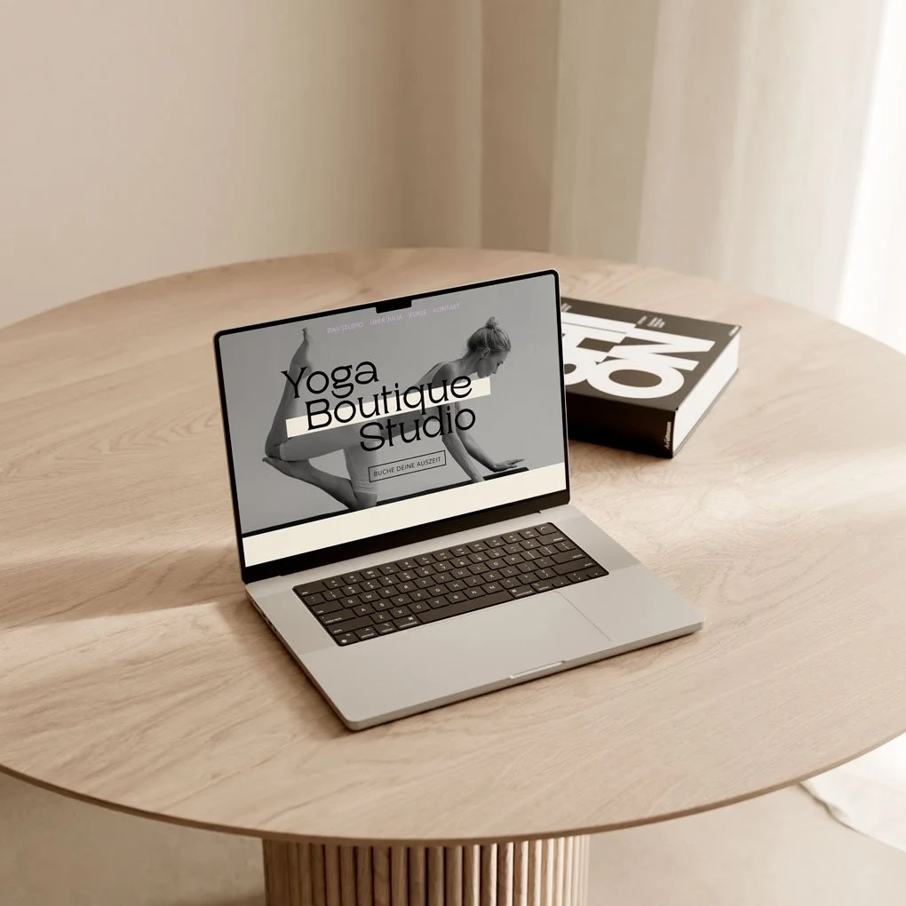 Laptop zeigt eine Webseite für ein Yoga-Boutique-Studio, auf einem runden Holztisch, daneben ein Buch.