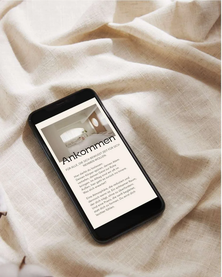Ein Smartphone liegt auf cremfarbenem Stoff. Auf dem Bildschirm ist eine Webseite mit dem Titel 'Ankommen' und einem Bild eines Schlafzimmers zu sehen.