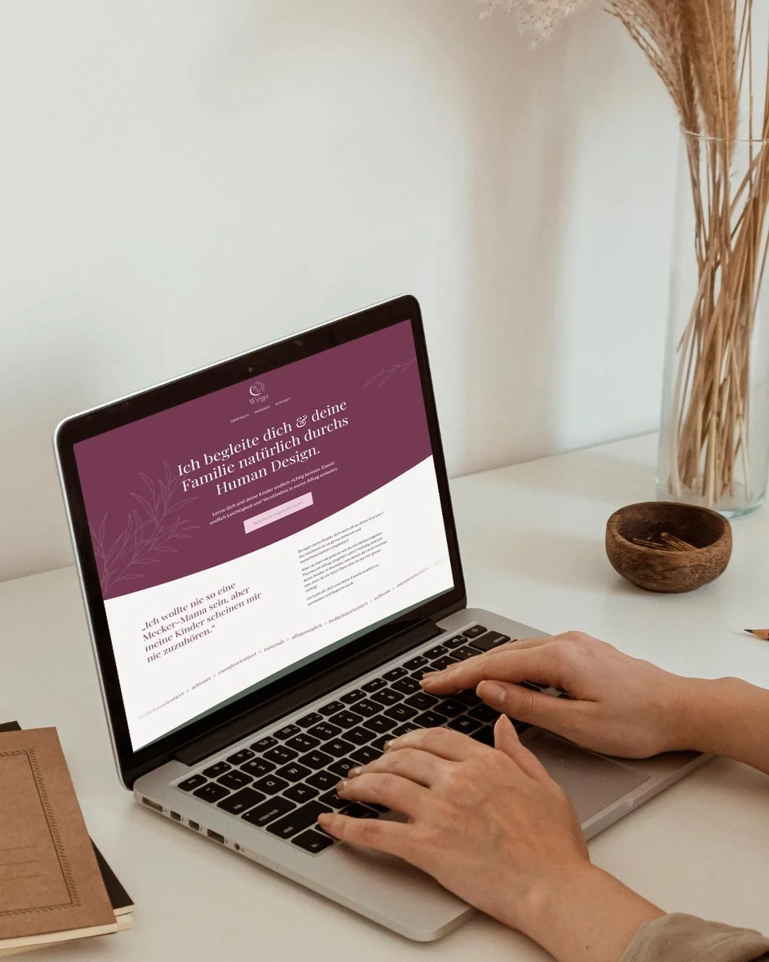 Person tippt auf einem MacBook-Laptop auf einem weißen Schreibtisch, neben einem braunen Notizbuch, einer Glaskaraffe mit Trockenblumen und einer kleinen Schale mit Gabeln.