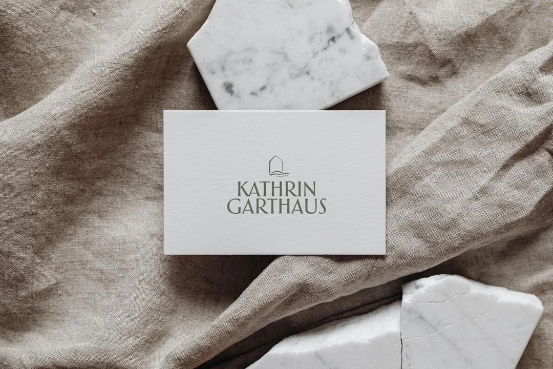 Visitenkarte mit dem Namen 'Kathrin Garthaus' auf einem beige Stoff, umgeben von weißen Marmorstücken.