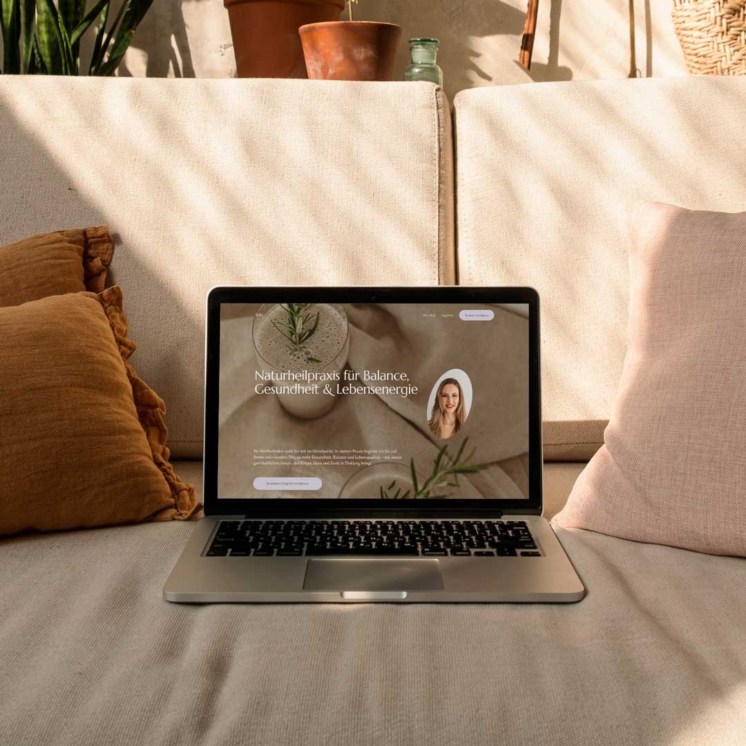 Ein Laptop liegt auf einem Sofa mit Kissen, zeigt eine Webseite für Naturheilpraxis. Im Hintergrund sind Pflanzen und Töpfe sichtbar.
