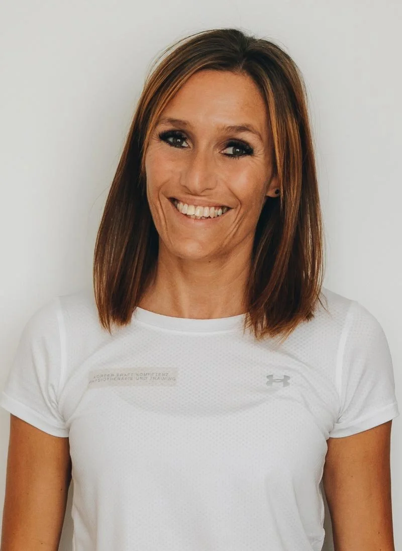 Eine lächelnde Frau mit braunen, geraden Haaren trägt ein weißes Sportshirt vor einem weißen Hintergrund.