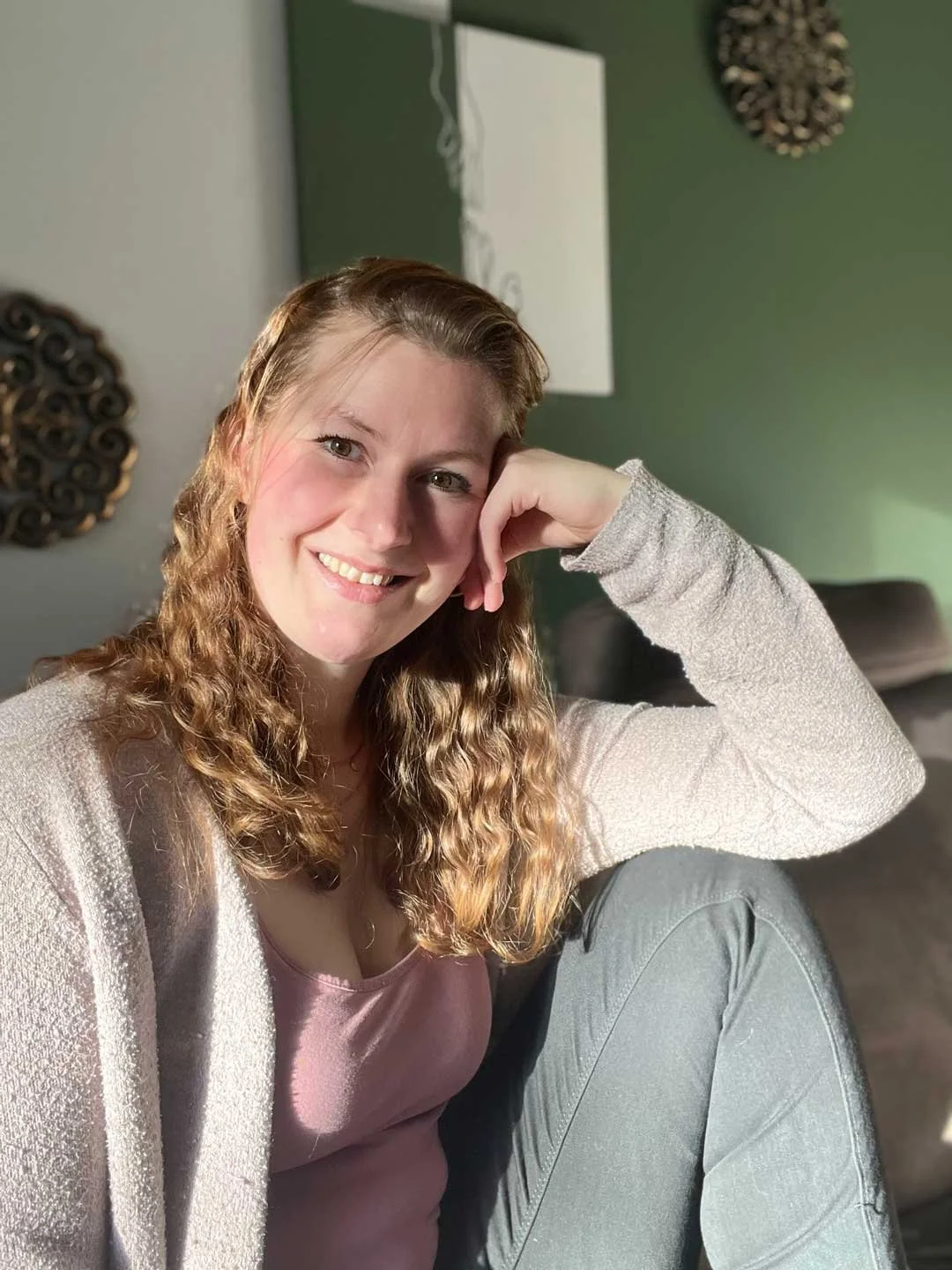Eine lachende junge Frau mit lockigem, hellbraunem Haar sitzt in einem Raum mit grüner und grauer Wand und modernen Wanddekorationen.