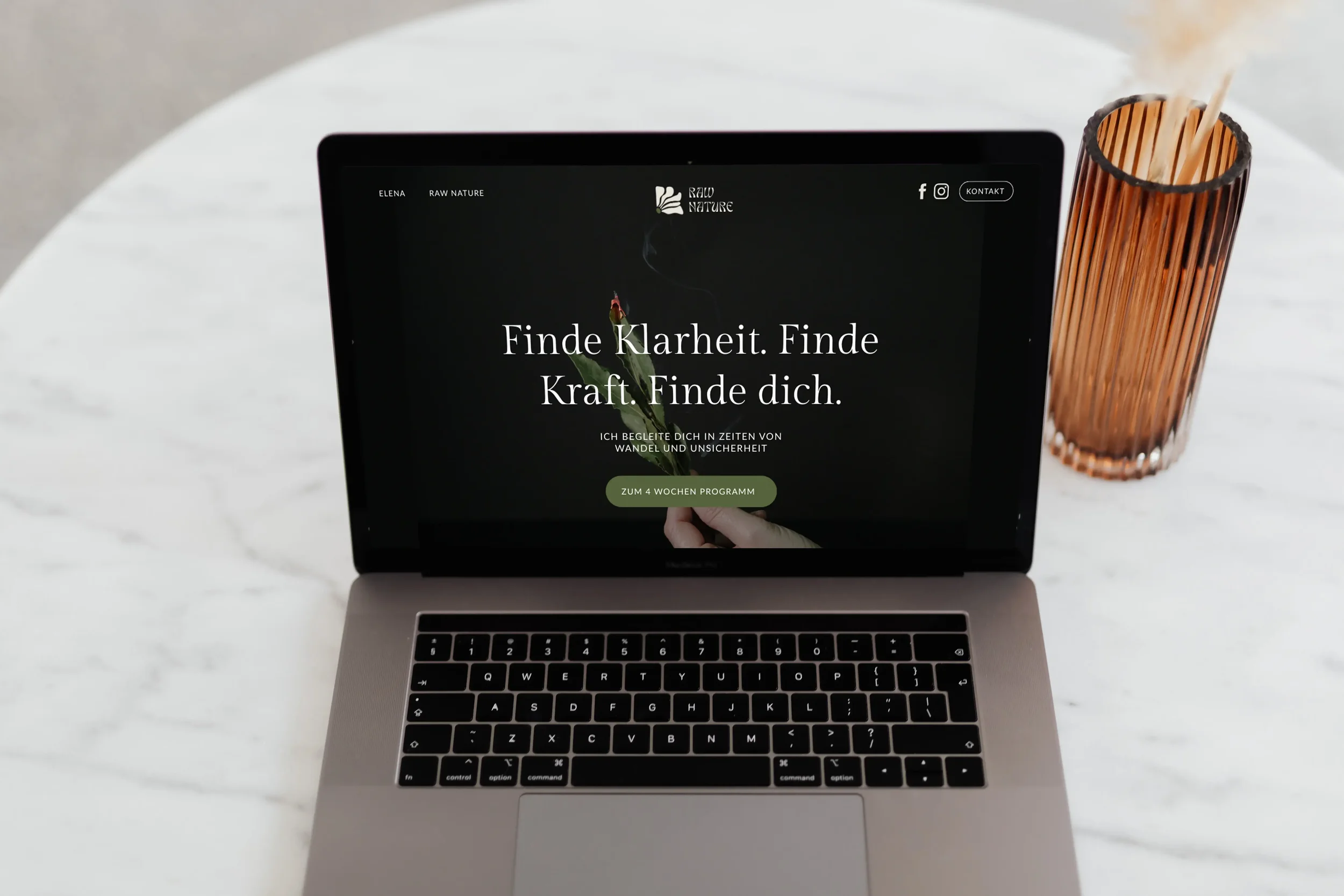 Offener Laptop auf weißem Marmor Tisch, der eine Webseite mit dunklem Hintergrund zeigt, die den Text 'Finde Klarheit. Finde Kraft. Finde dich.' enthält. Neben dem Laptop steht ein roségoldener Stiftbecher, in dem Stifte und eine Kerze stecken.