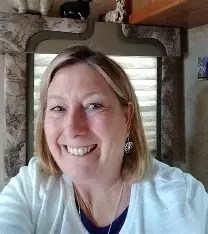 Smiling woman with shoulder-length blonde hair and earrings, indoors with a window and stone wall in the background.