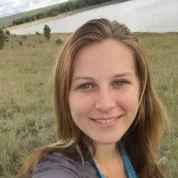A young woman with shoulder-length hair smiling outdoors near a body of water and grassy landscape.