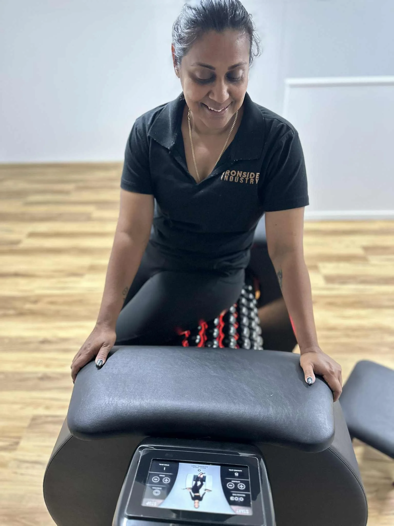 Woman in black polo shirt kneeling on a lymphatic roller machine, smiling.