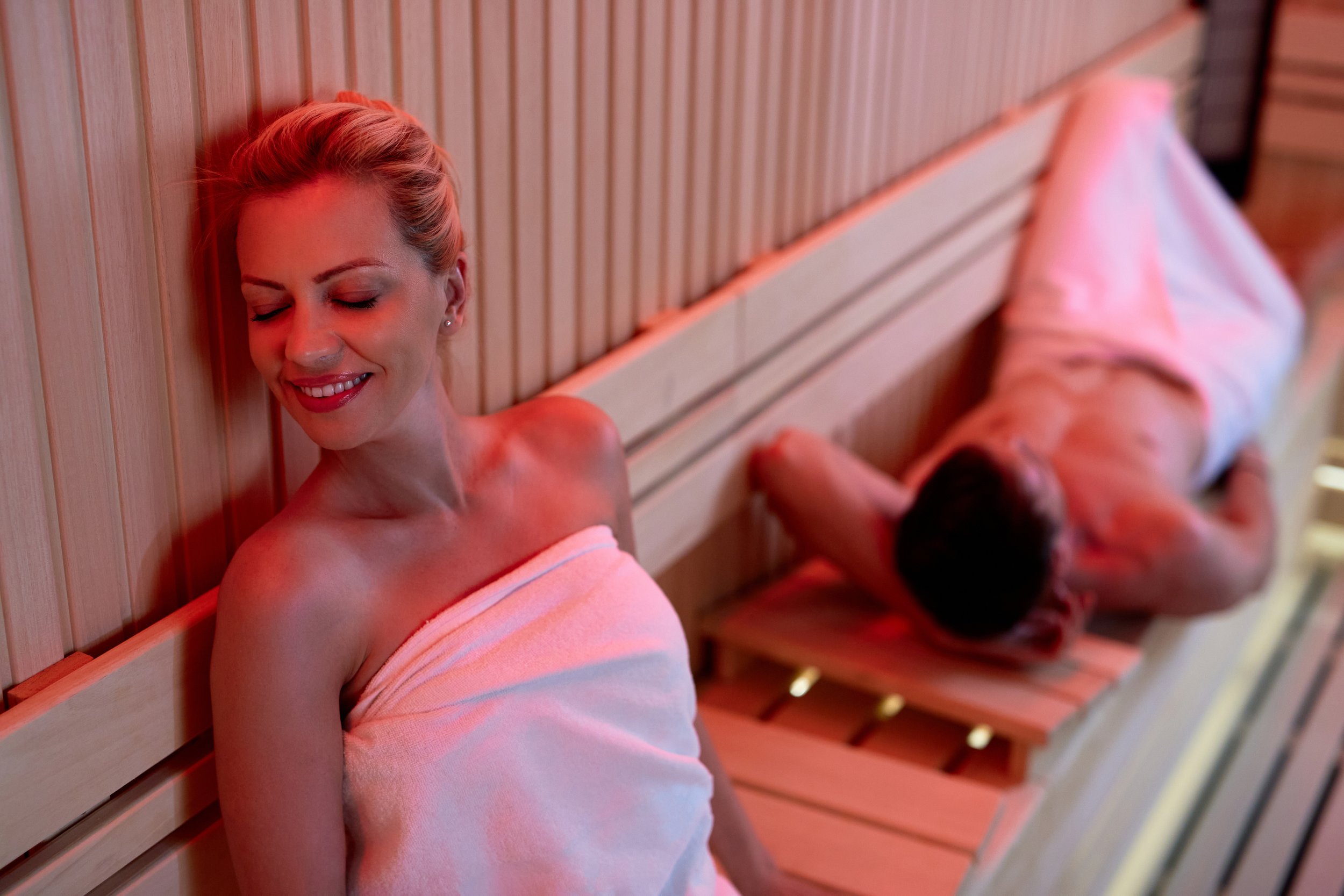 A woman smiling with eyes closed, wrapped in a towel, sitting in a sauna. A man is lying on a bench in the background, also wrapped in a towel, with his face turned away.