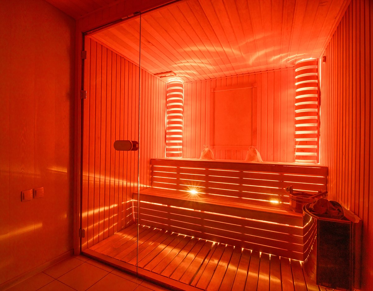 A modern infrared sauna room with wooden walls and benches, illuminated with red lighting, enclosed by glass doors.