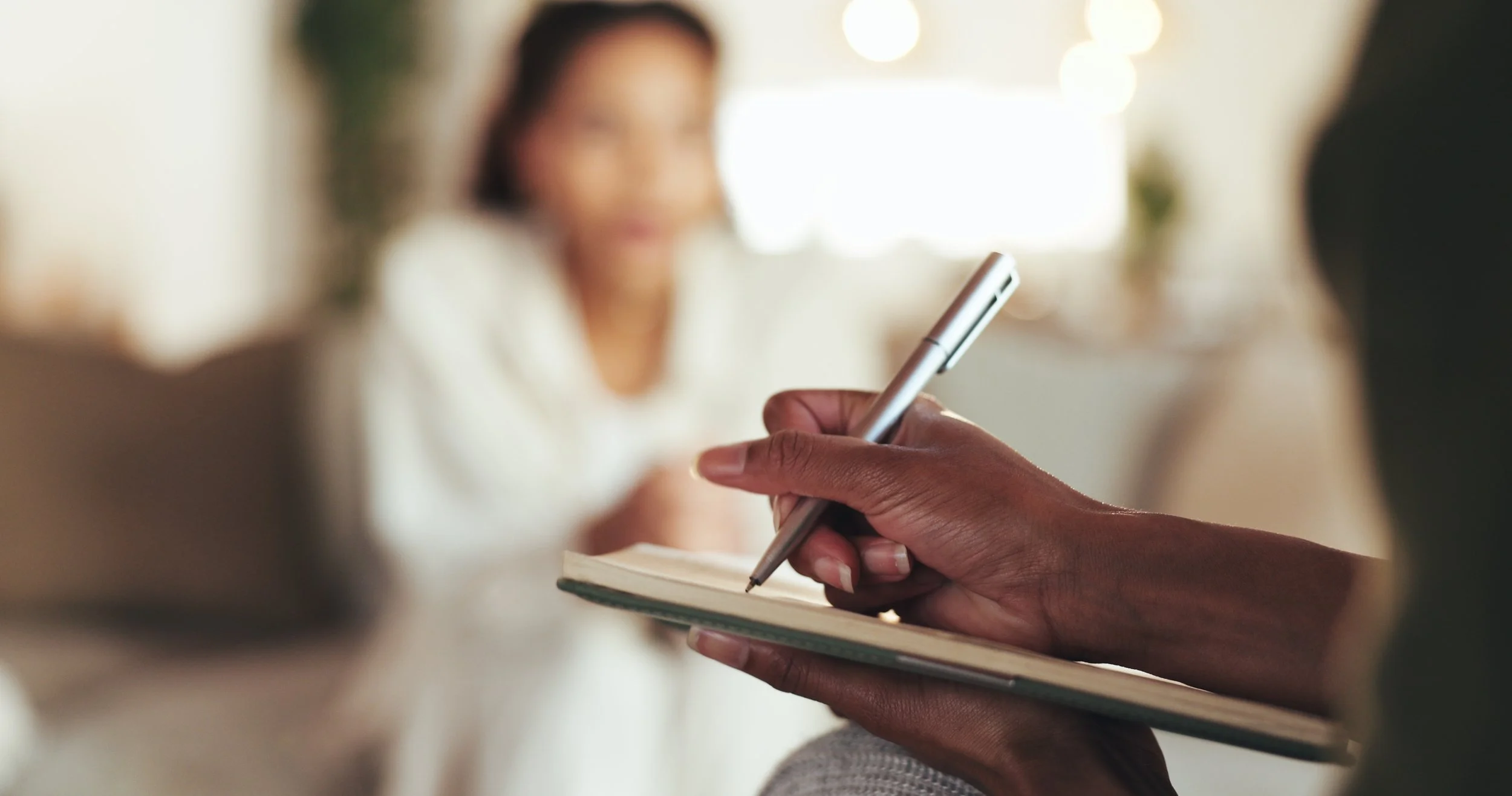 Close-up of a person taking notes in a notebook with a silver pen, with a blurred woman in the background.