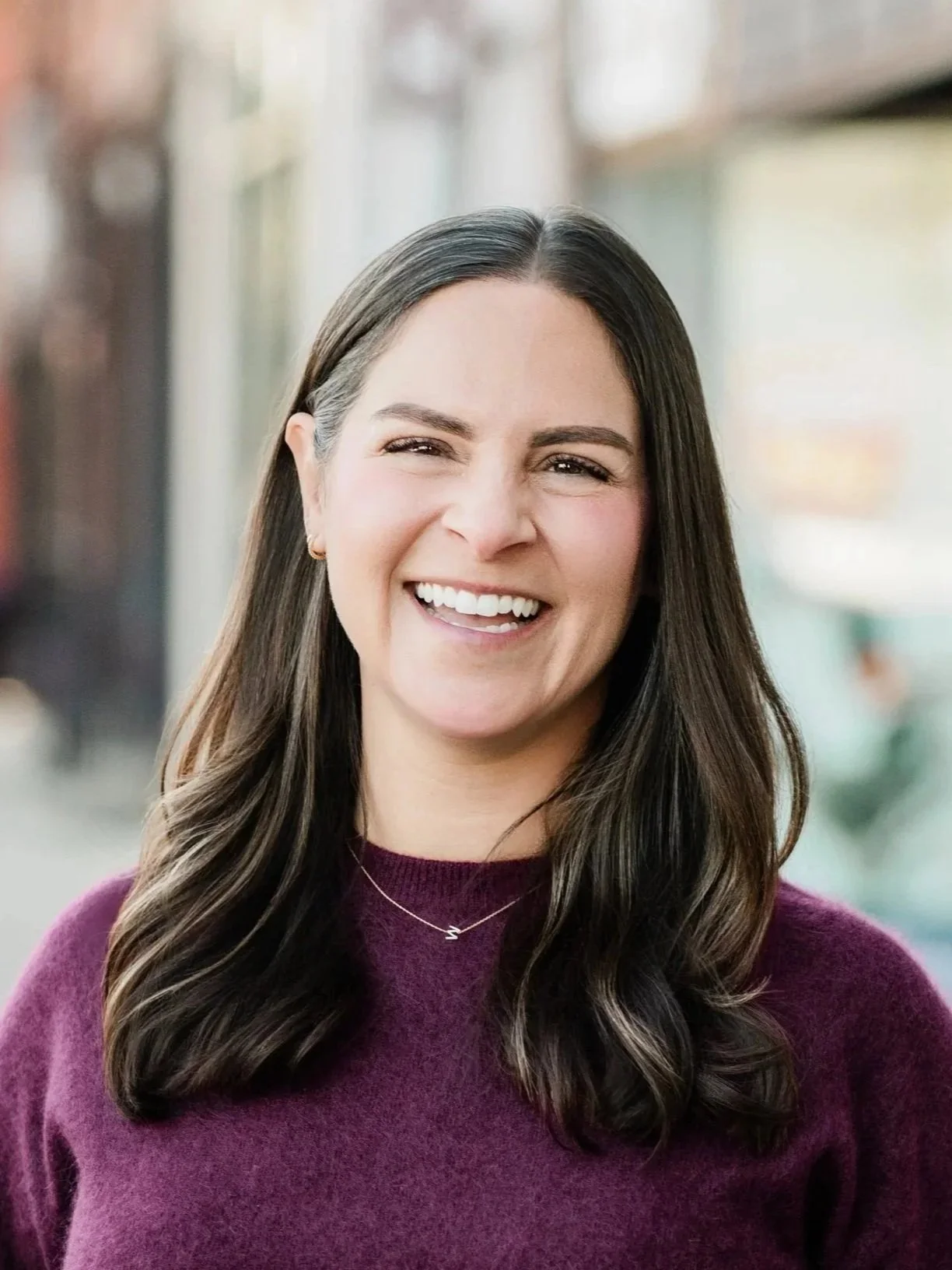 A woman with long dark hair smiling outdoors, wearing a purple sweater and a delicate necklace.