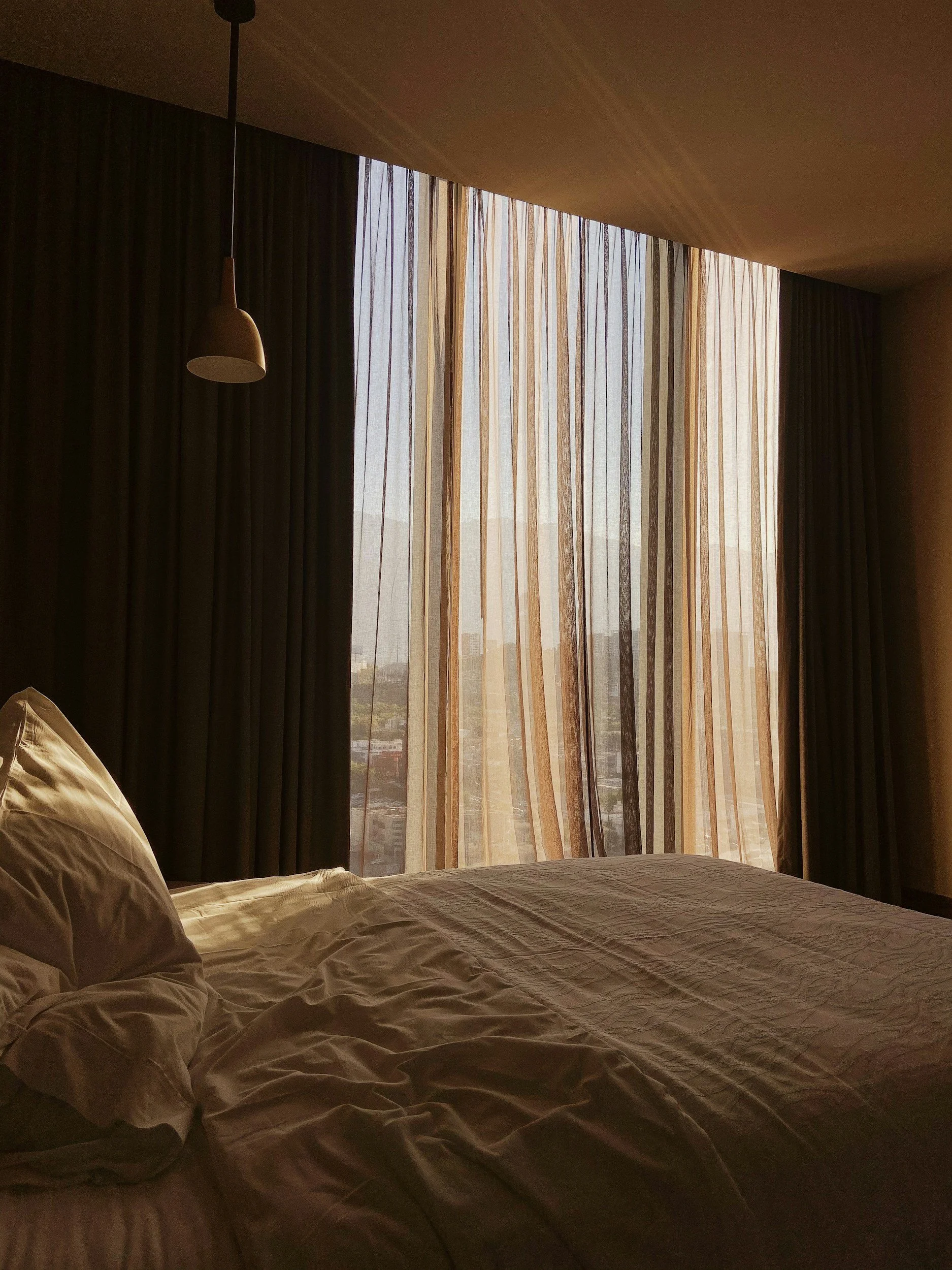 Sunlit hotel room with unmade bed, sheer curtains, and blackout curtains, showing city skyline outside.
