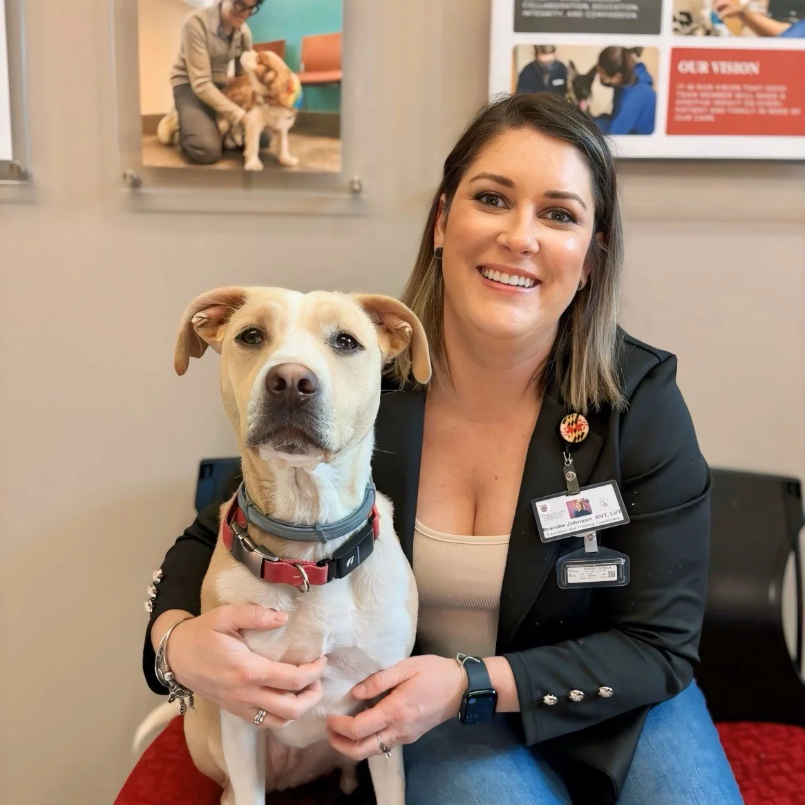 A woman smiling and holding a light-colored dog with a red collar inside a veterinary office. The woman is wearing a black blazer with a name tag and a watch, and has short brown hair. In the background, there are posters and another photo of a person with a dog.