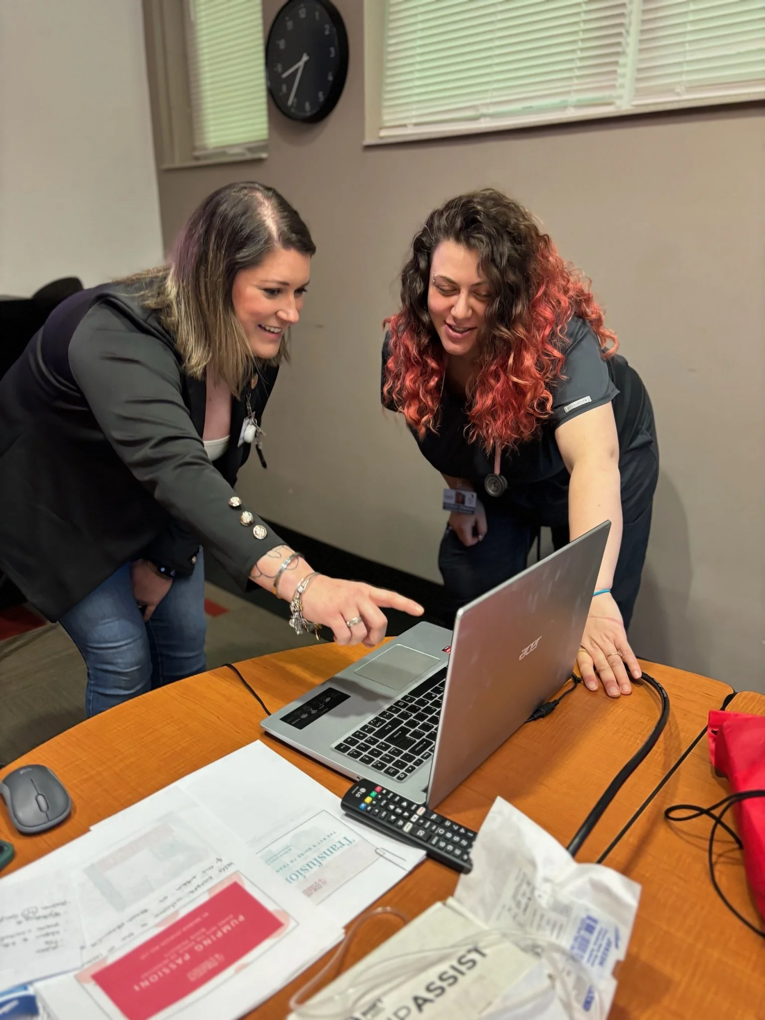 Two women, one with curly hair and another with straight hair, are looking at a laptop screen together in an office, smiling and pointing. There are papers, a remote, a mouse, and a red bag on the table.