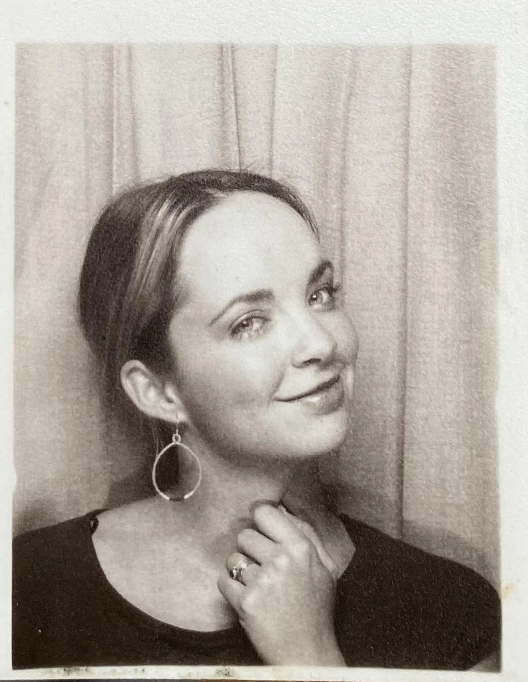 A black and white photo of a woman with short hair, wearing a dark top, hoop earrings, and a ring on her hand, standing in front of a curtain and smiling at the camera.