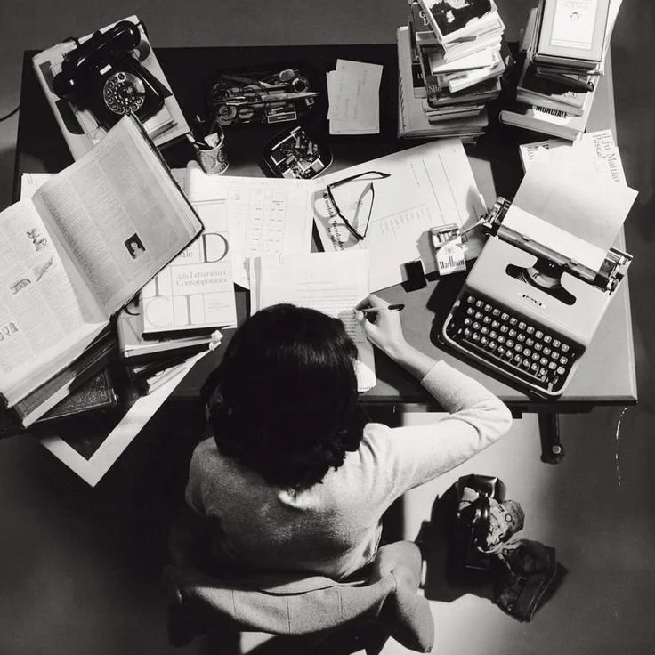 Overhead view of a woman working at a cluttered desk with books, papers, a typewriter, and office supplies.