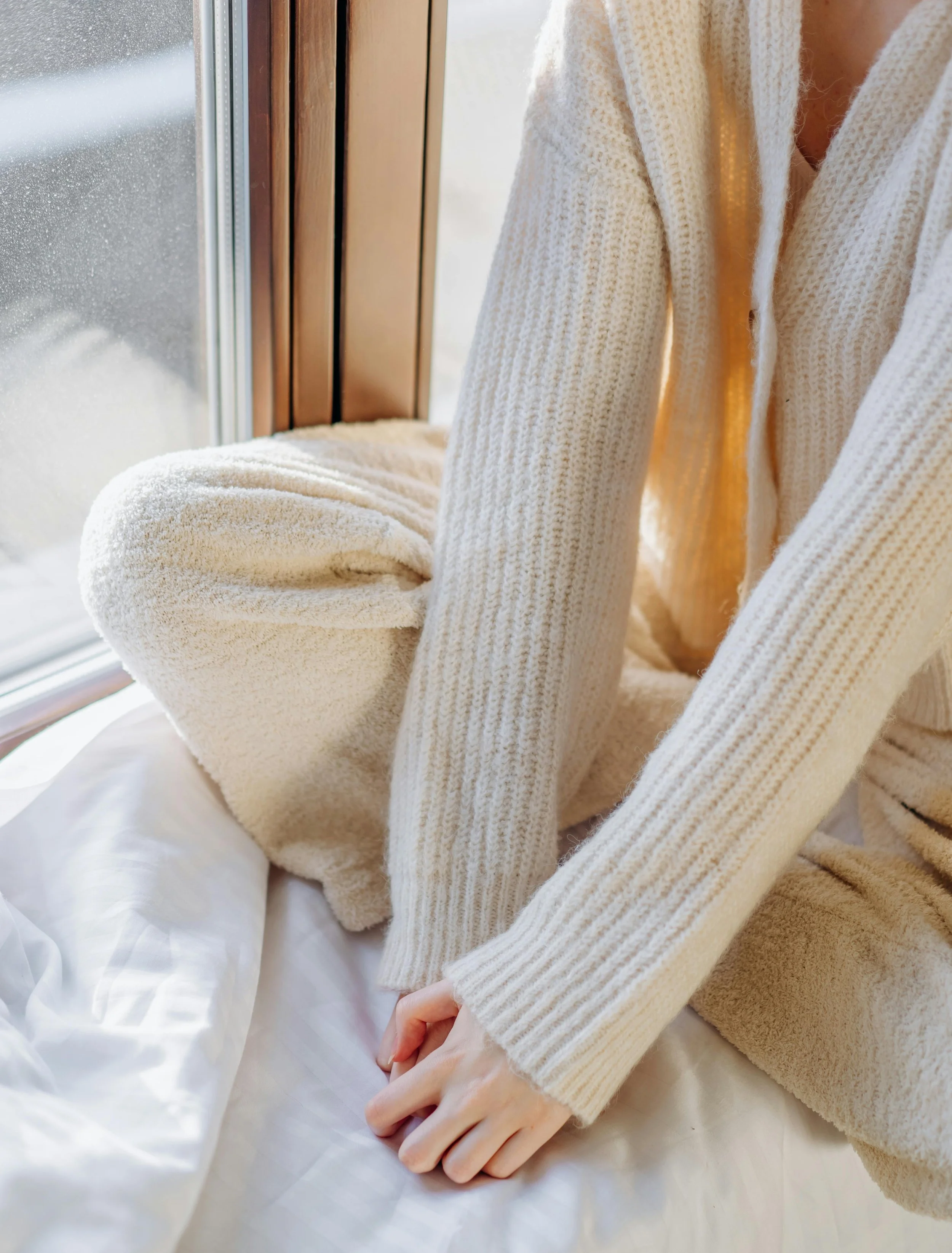 Person sitting by a window in cozy cream sweater and pants, with sunlight streaming in.