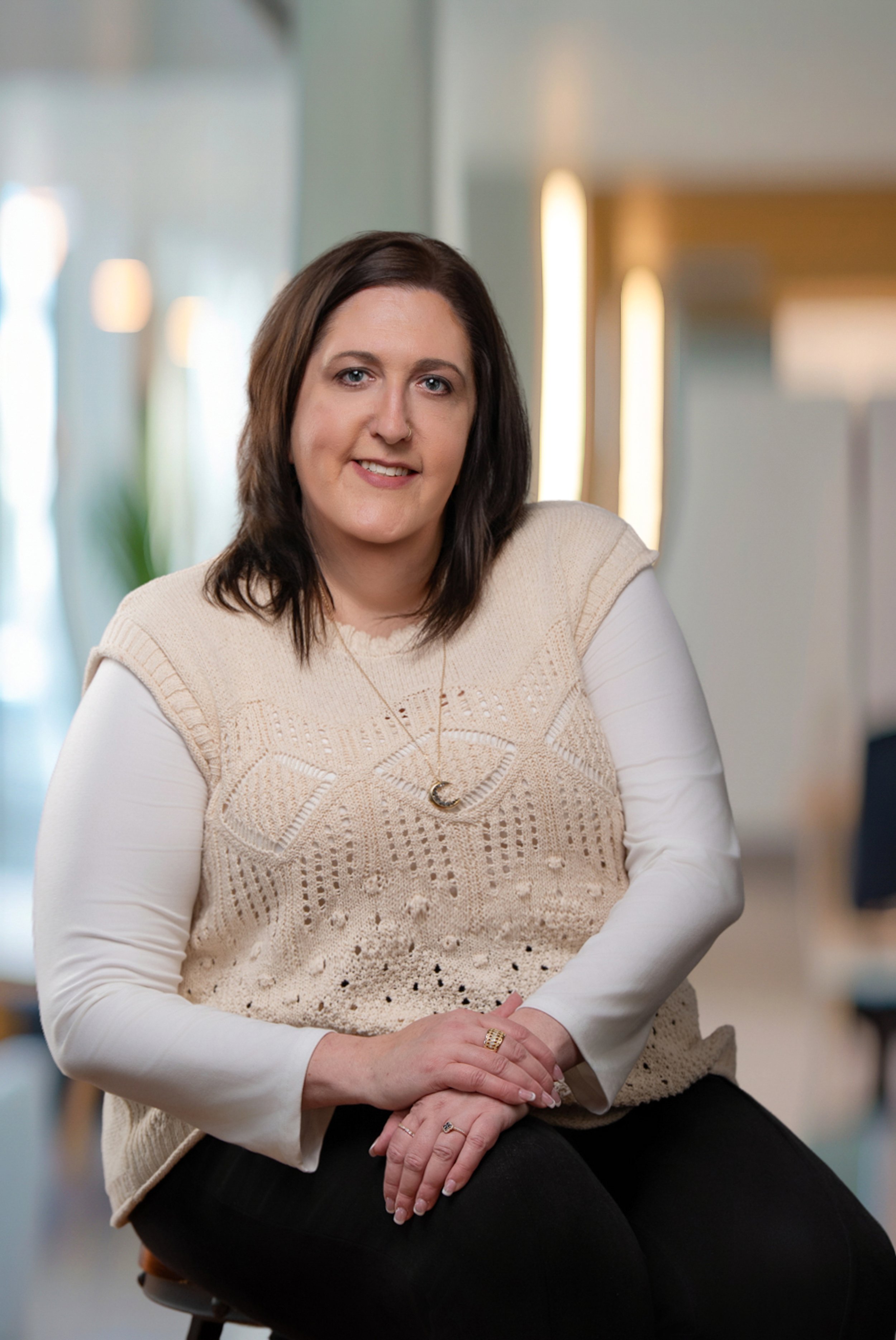 A woman with shoulder-length dark brown hair, wearing a beige knitted vest over a white long-sleeve shirt, sitting indoors with hands resting on her lap, smiling at the camera.