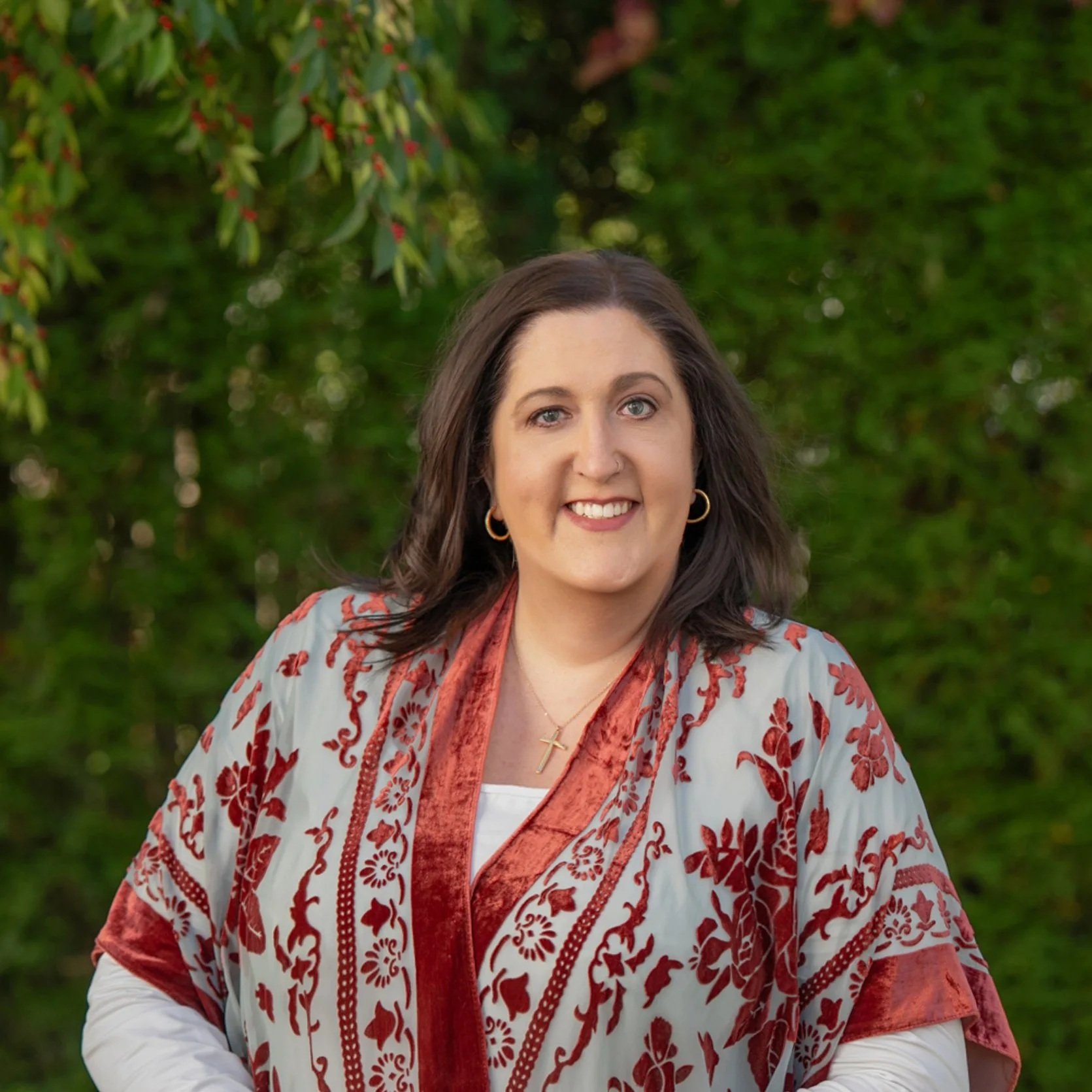 A woman with dark brown hair, blue eyes, and a bright smile standing outdoors in front of greenery, wearing a patterned shawl with red and white colors, gold hoop earrings, a gold cross necklace, and a white top.