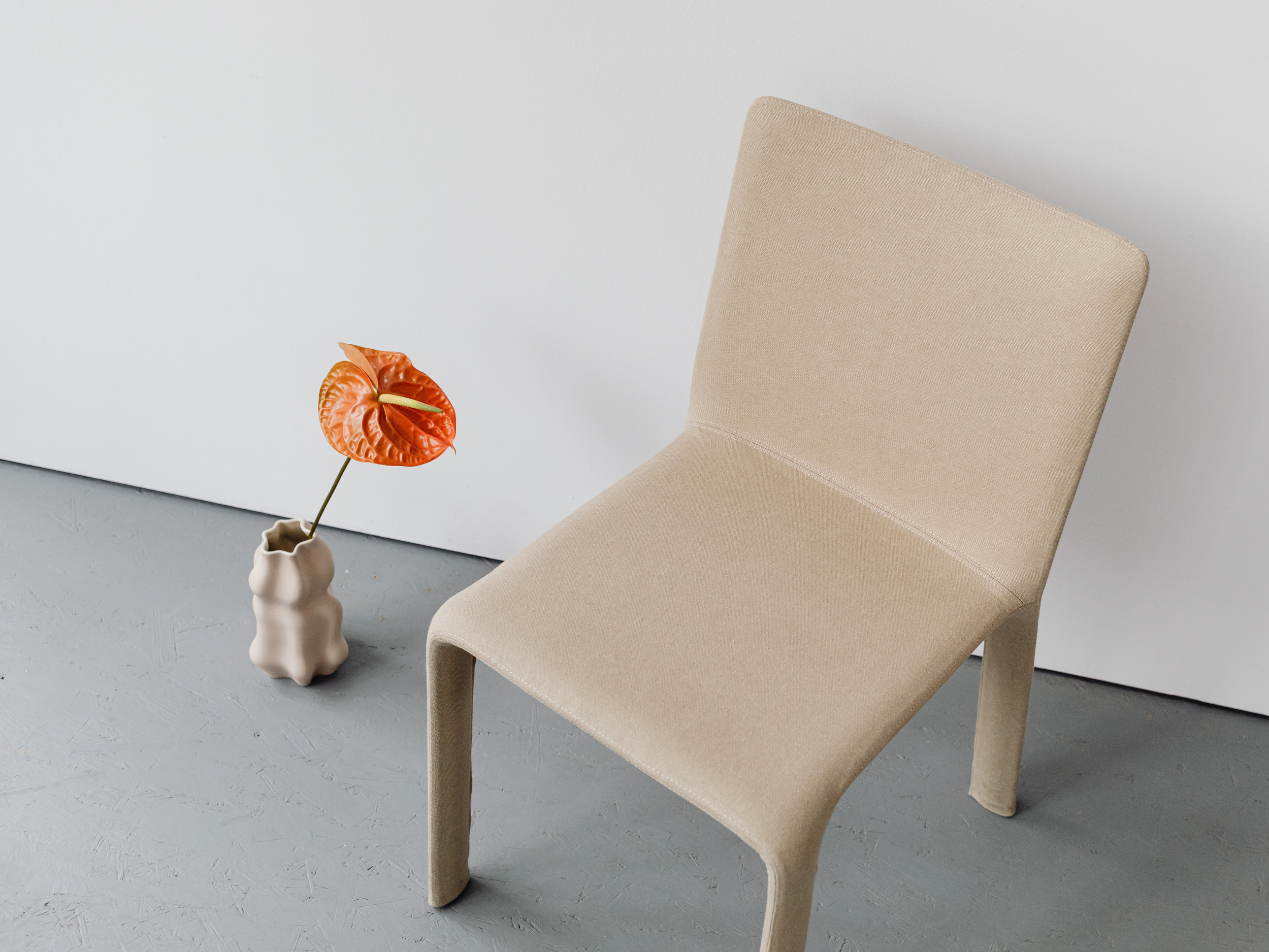 A beige upholstered chair with a high back is placed next to a small beige vase with a textured surface, holding an orange and green anthurium flower, against a gray floor and white wall background.