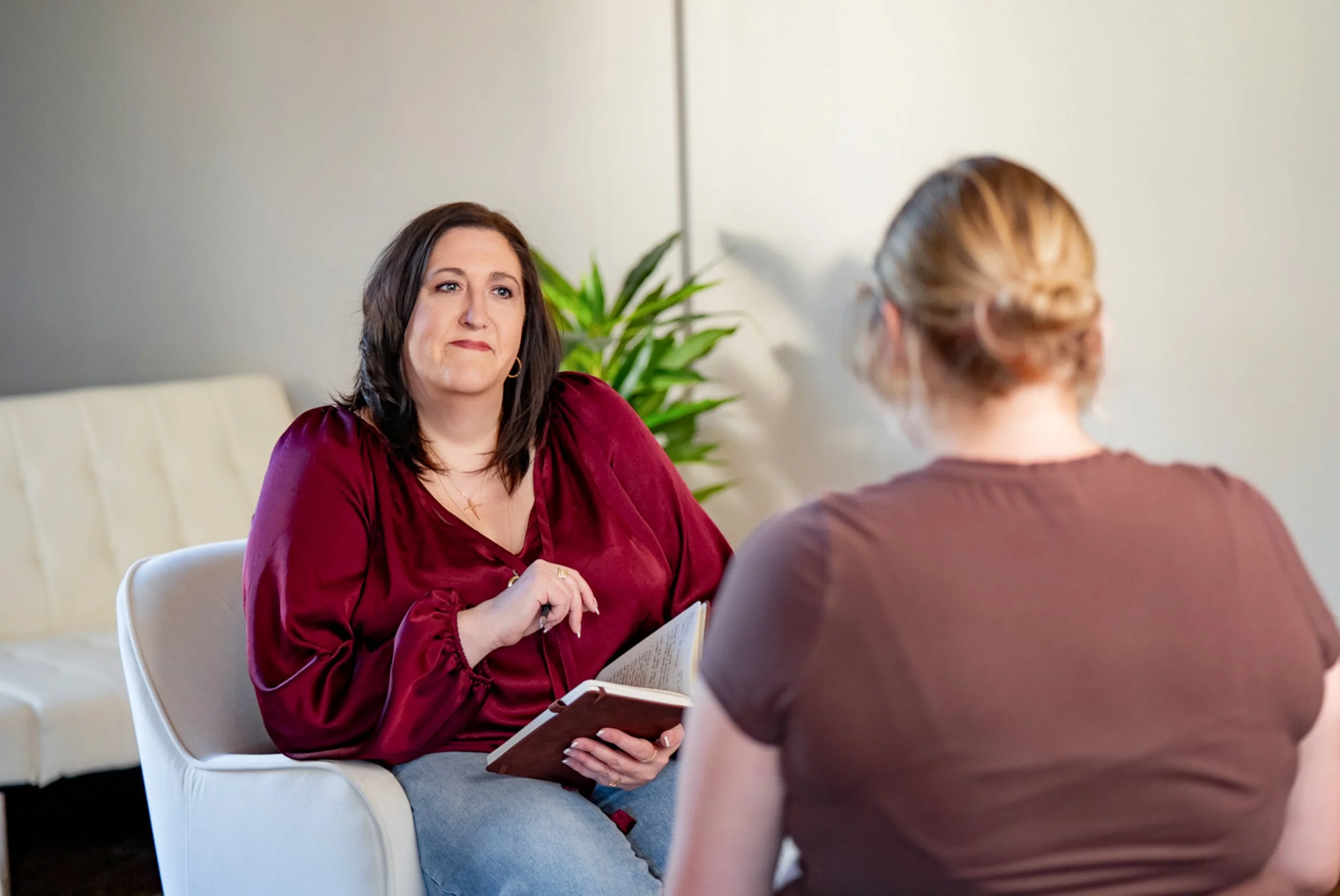 A woman wearing a maroon blouse and holding a book is sitting in a white chair, facing another woman with blonde hair in a bun, who is seen from behind. They are in a room with a gray wall and a green potted plant.