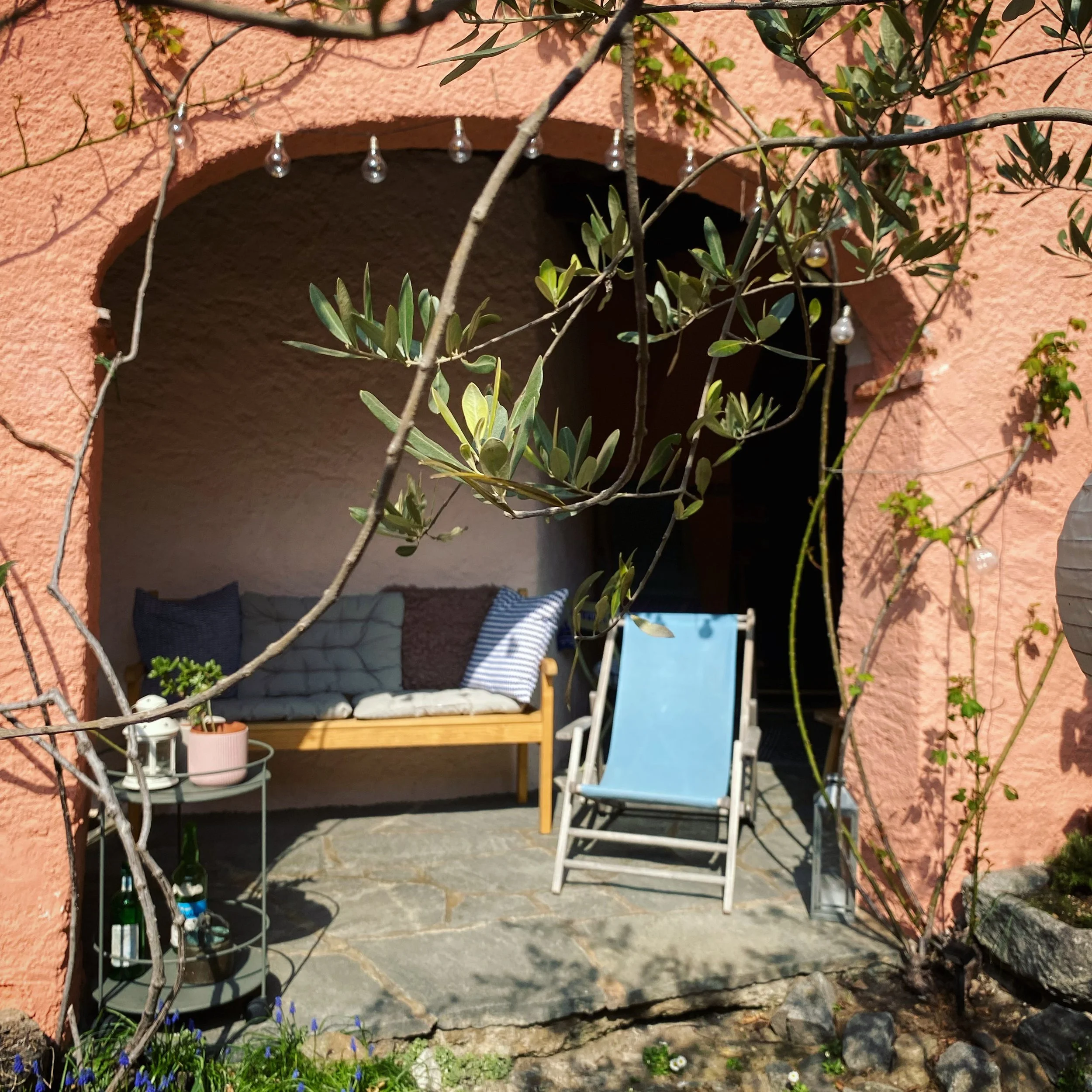 Veranda con panca di legno, cuscini, sedia blu pieghevole e tavolino con piante, bottiglie e lanterna, circondata da piante e decorazioni luminose