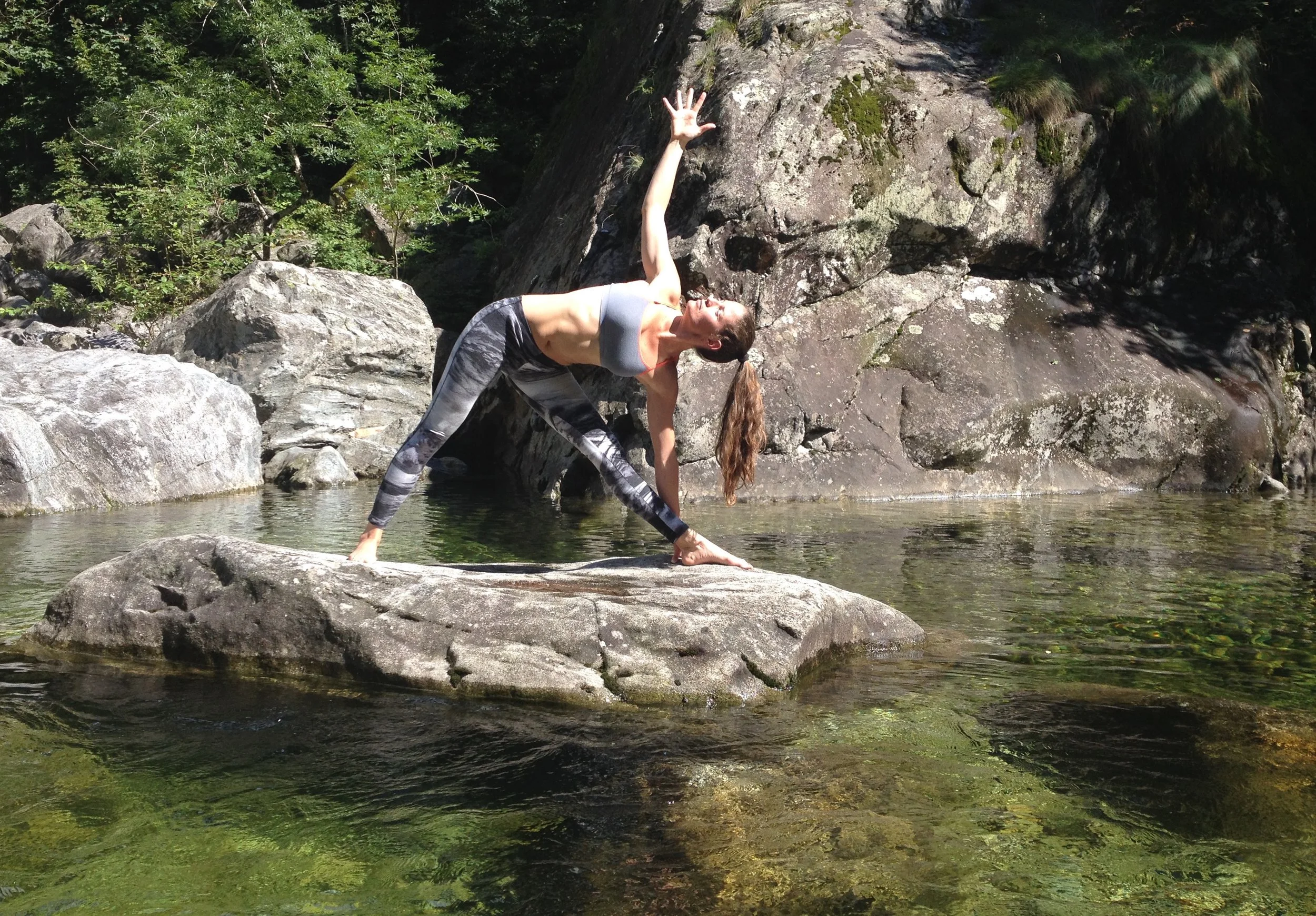 Donna che fa yoga su una grande roccia in un fiume, circondata da natura verde, in una posa di stretching con una mano rivolta verso il cielo.