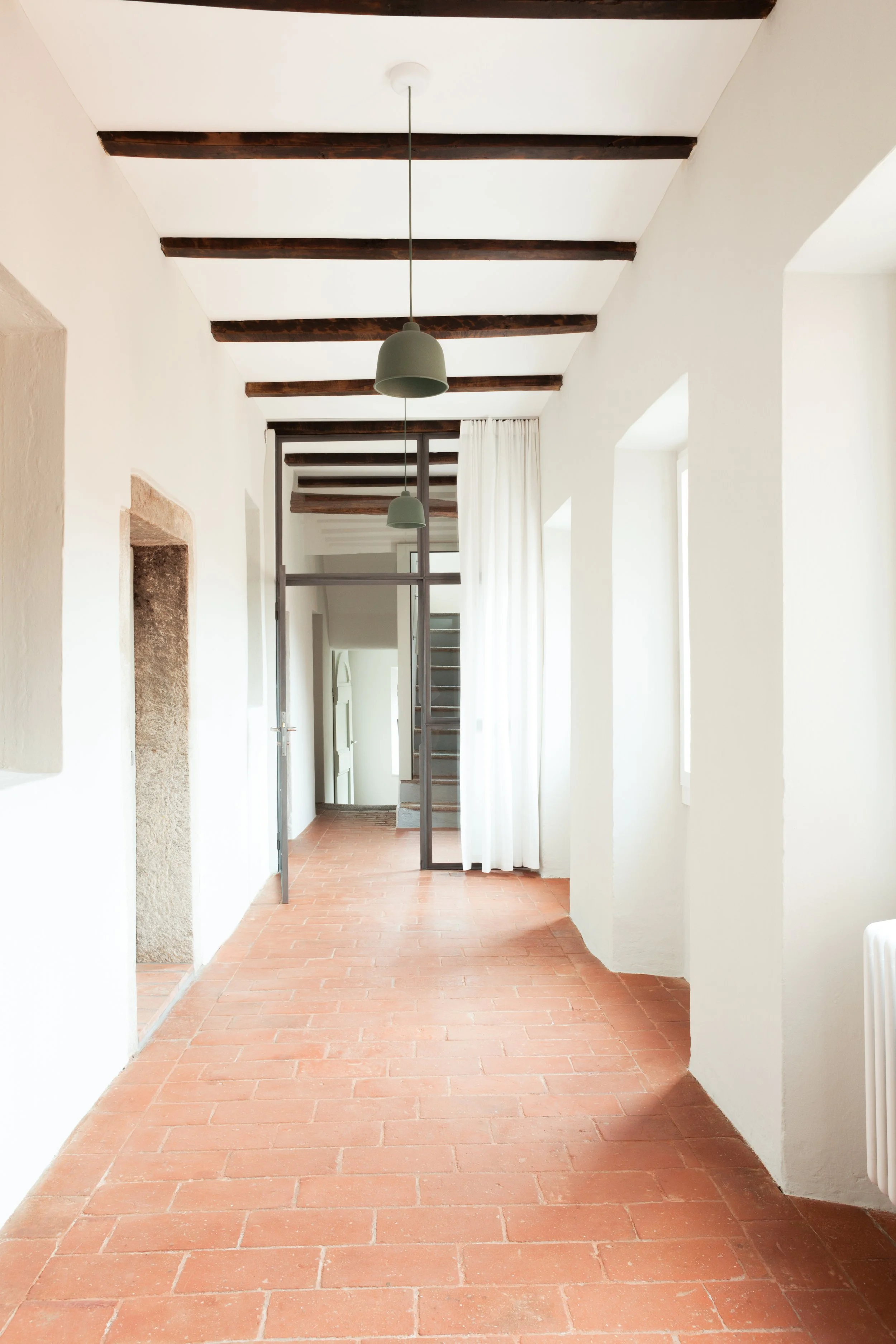 Interno di una casa con pareti bianche, soffitto con travi di legno, pavimento in cotto, finestre con tende bianche, lampade a pendant verde chiaro, e una porta di vetro con una scala visibile attraverso.