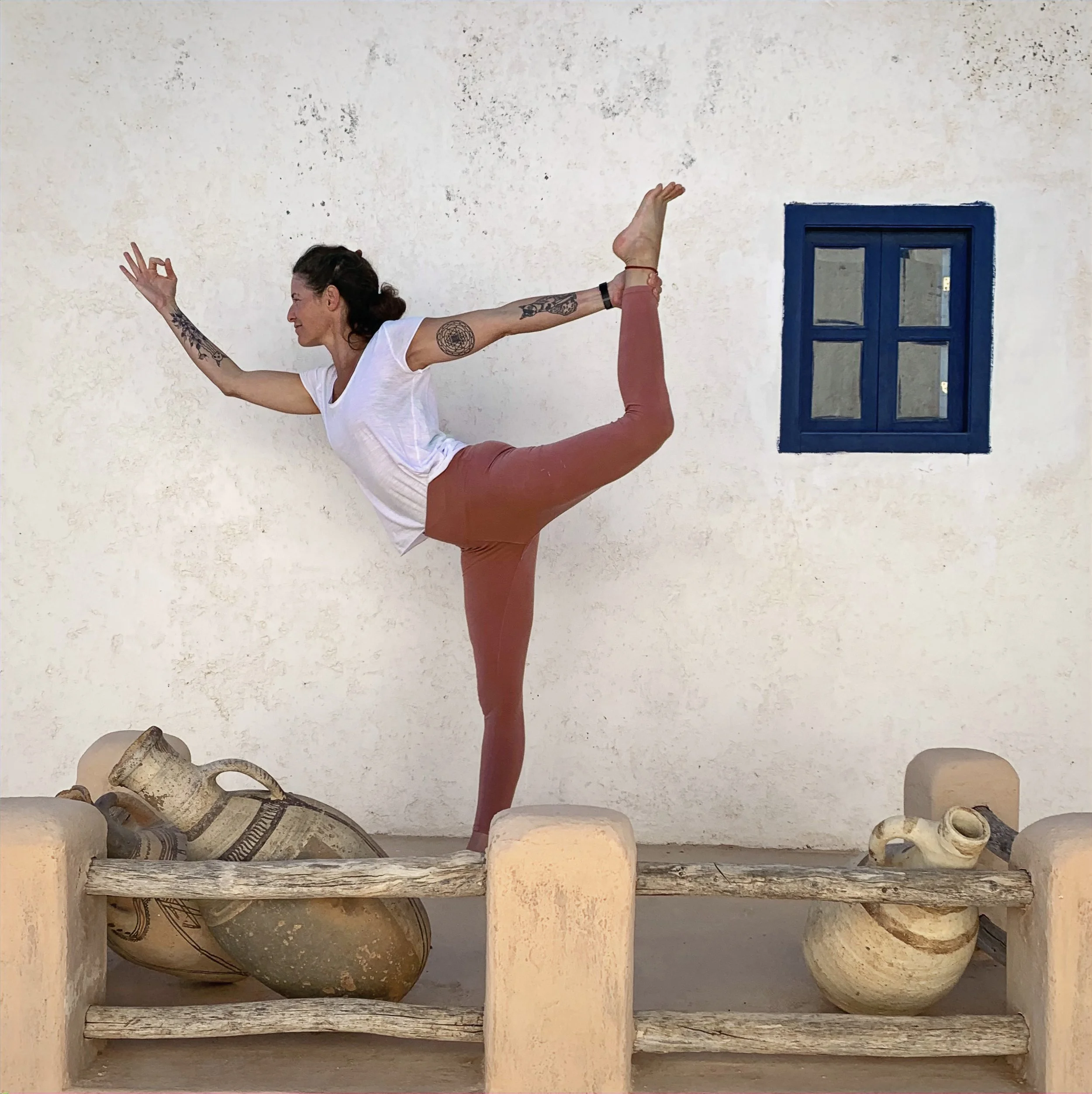Una donna in posa yoga in equilibrio su una gamba, con l'altra gamba sollevata dietro di lei e afferrata con una mano, davanti a un muro bianco con una piccola finestra blu.
