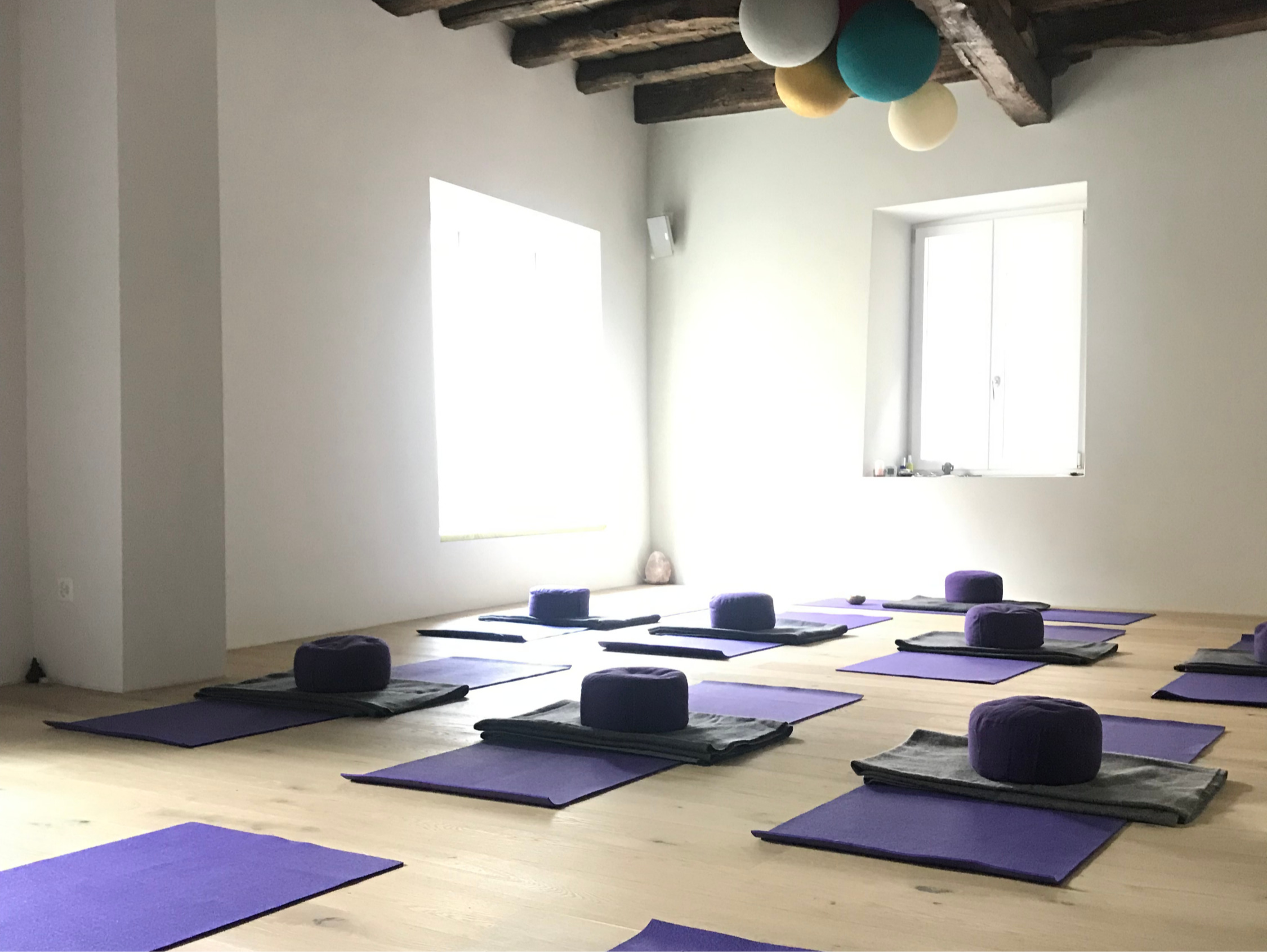 Studio di yoga con tappetini viola e cuscini rotondi in una stanza con pareti bianche, soffitto con travi di legno e finestre che lasciano entrare luce naturale.
