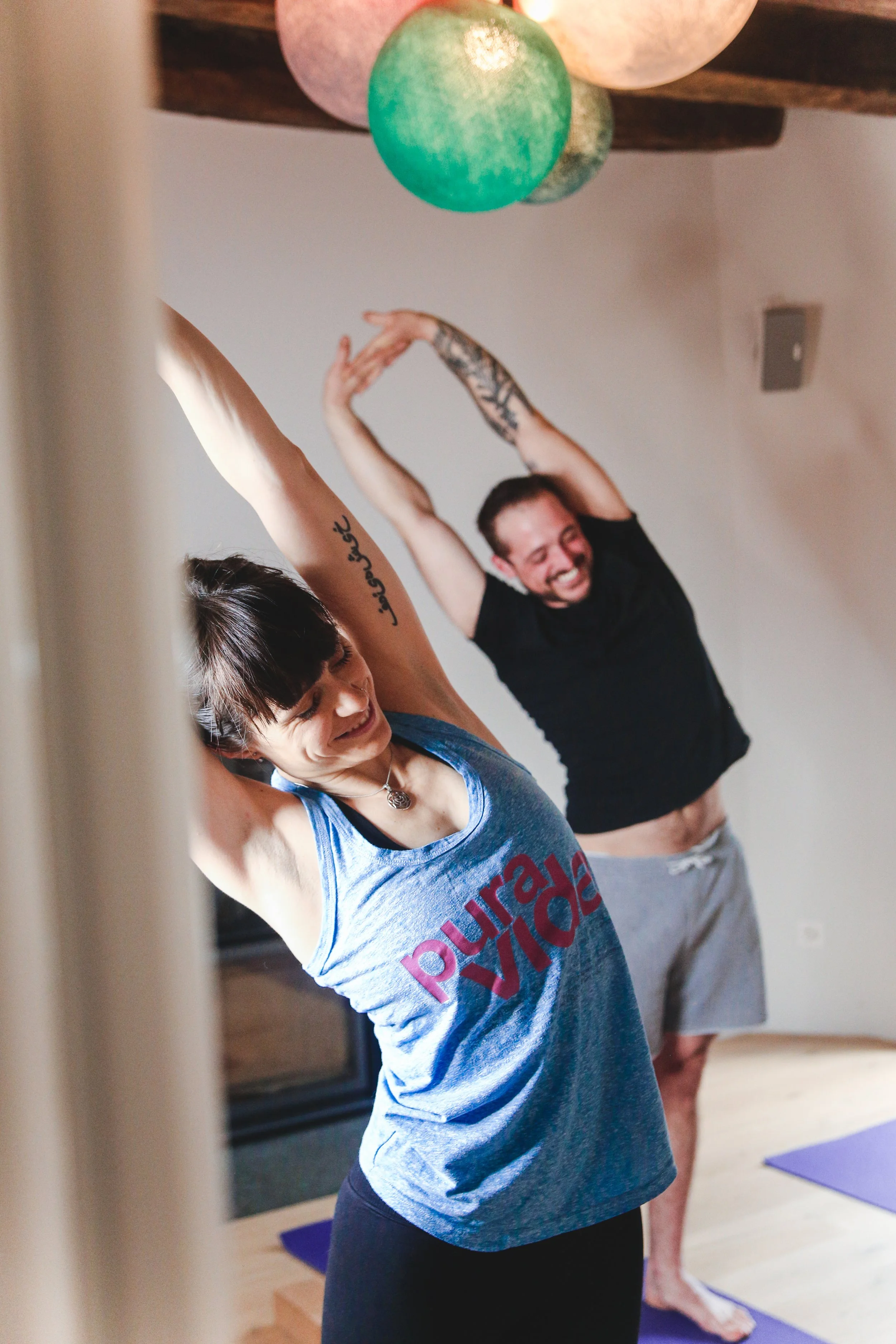 Una donna e un uomo stanno facendo stretching insieme in una stanza, probabilmente durante una lezione di yoga o pilates, con sfondo semplice e luci naturali.