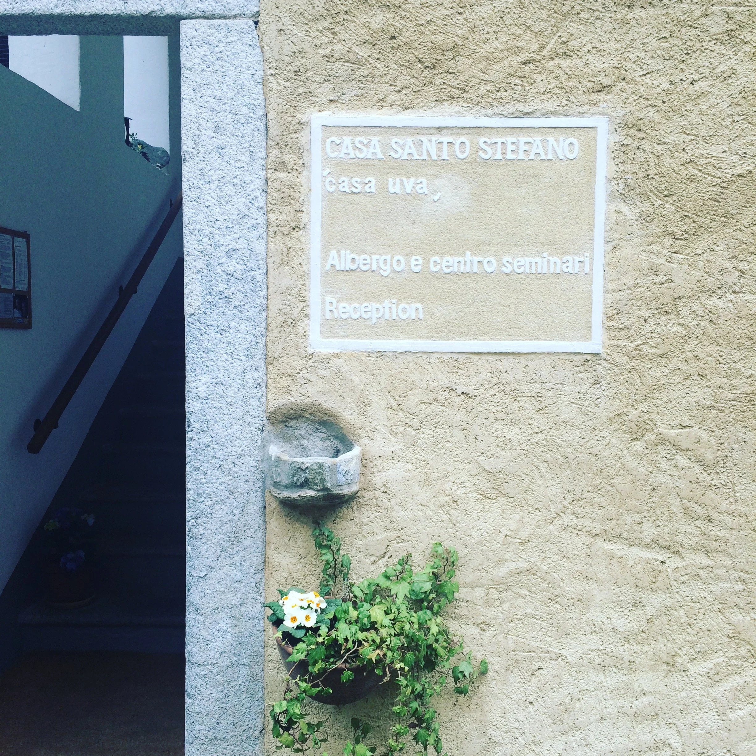Segnale di un edificio con un'edicola con fiori e una parete in pietra, scritto 'Casa Santo Stefano' con ulteriori informazioni in italiano e inglese.