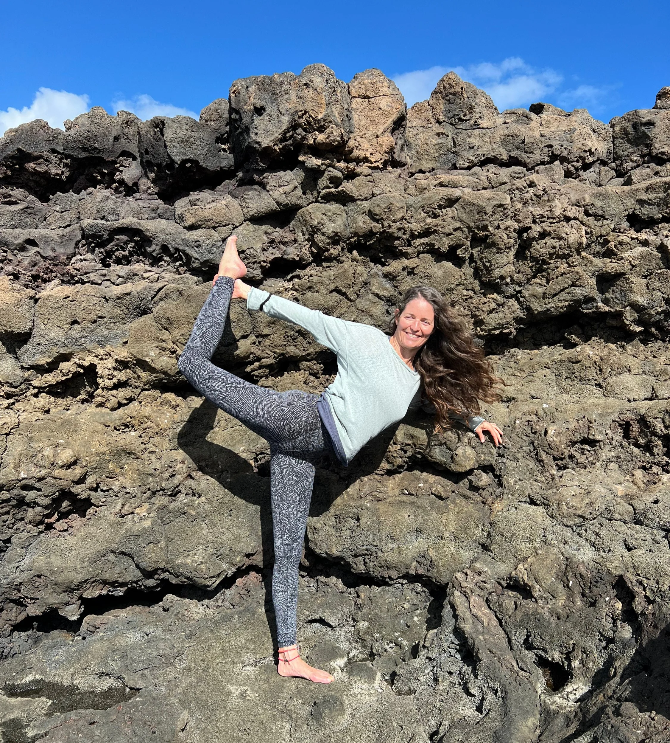 Una donna che fa yoga in posizione di equilibrio su un lato di un muro di rocce naturali, sorridendo sotto un cielo sereno.