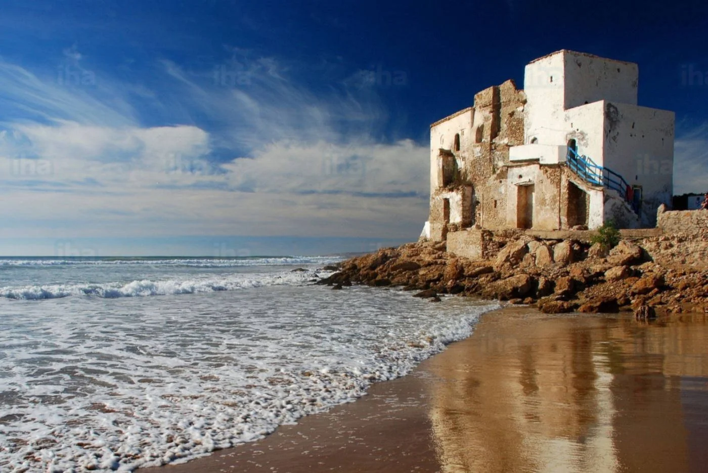 Un edificio antico sulla riva del mare, con onde che si infrangono sulla spiaggia di sabbia, sotto un cielo blu con nuvole sparse.