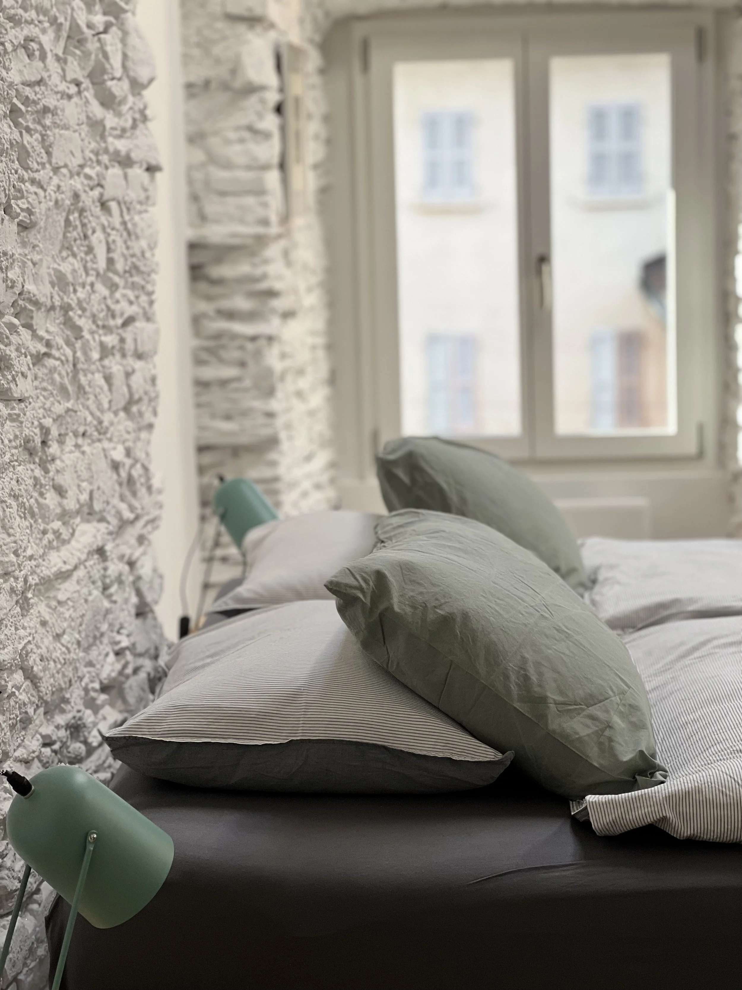 Letto con cuscini verdi e lenzuola con riga sottile, accanto a una lampada da tavolo verde, in una stanza con pareti in pietra bianca e finestra con vista su edifici adiacenti