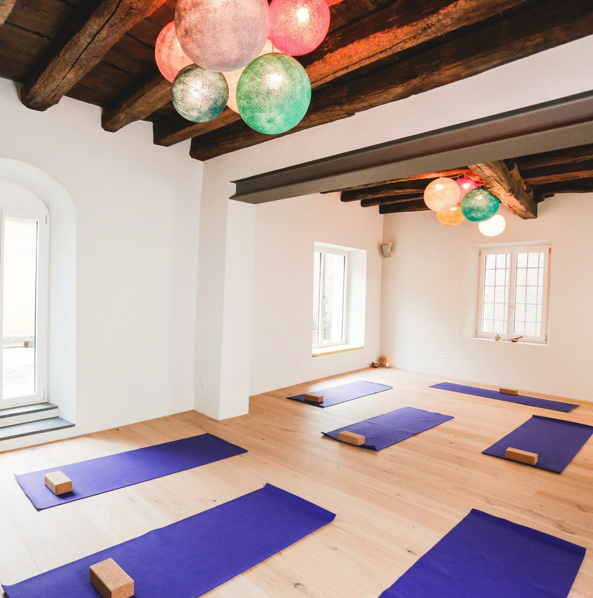 Stanza semplice con pareti bianche, soffitto con travi di legno esposte e lampadari colorati. Sei tappetini da yoga blu disposti sul pavimento di legno, ognuno con un blocco di supporto sotto.