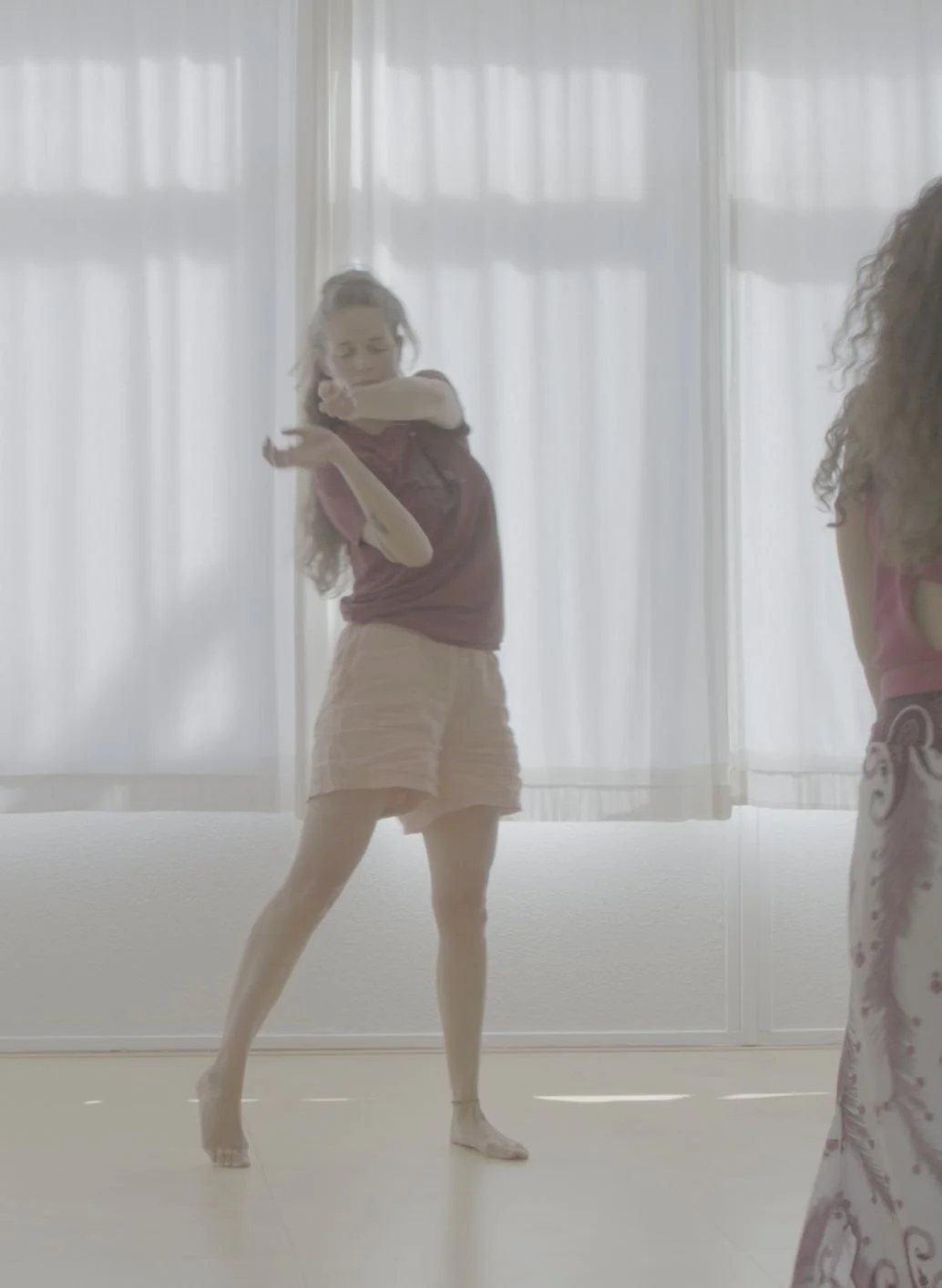 Donna che danza in una stanza con tendaggi bianchi, indossa una maglietta viola e pantaloncini beige, con i capelli ondulati e sciolti.