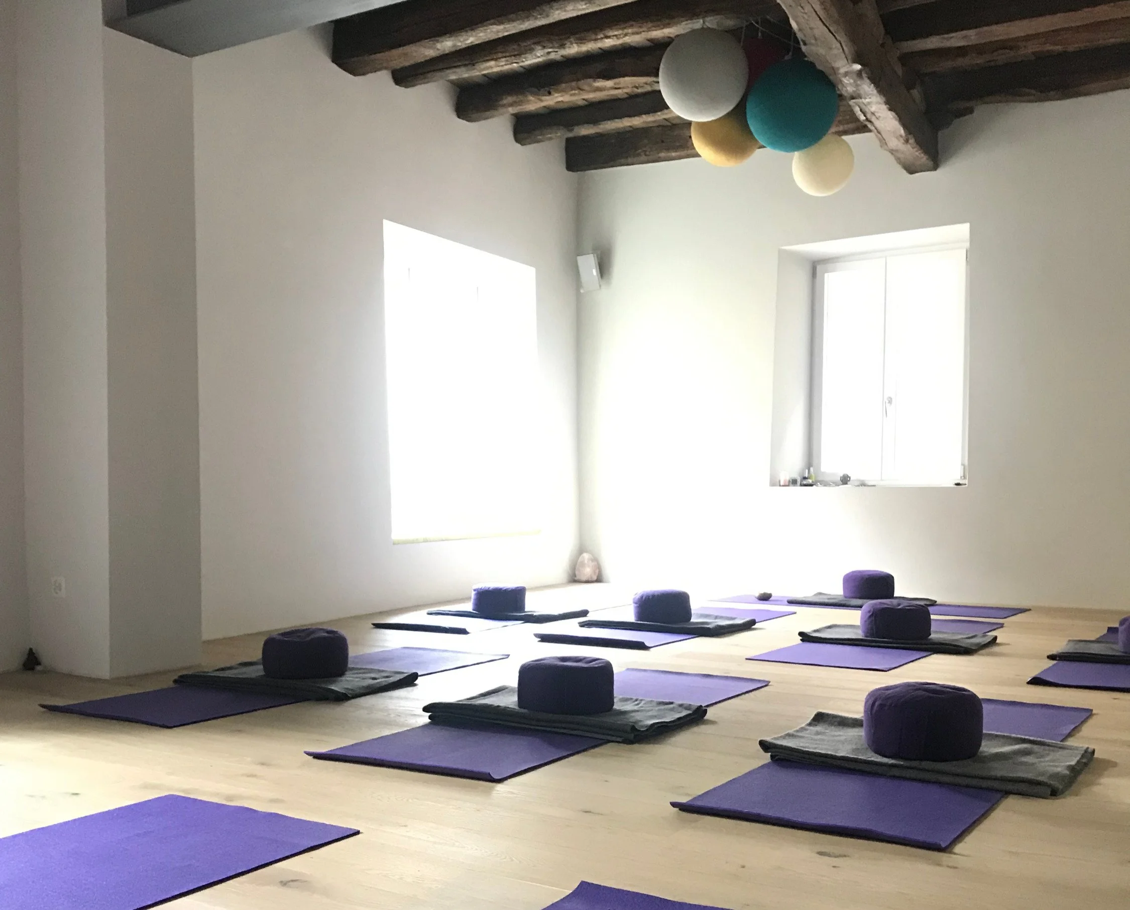 Stanza luminosa con tappetini da yoga viola, cuscini rotondi viola e tie-dye in stoffa grigia, alcune pareti con finestre e soffitto con travi di legno.