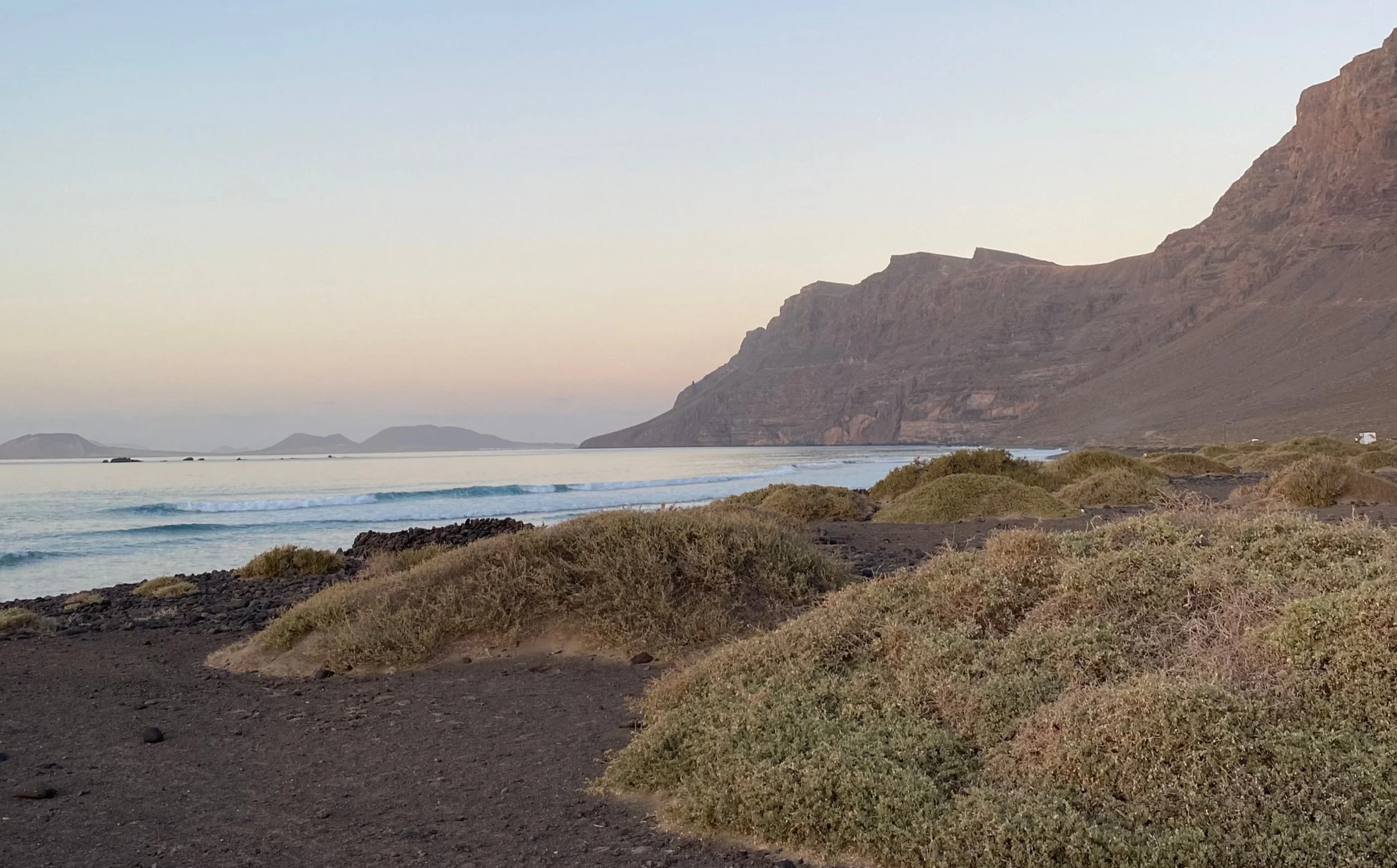 Spiaggia vulcanica con cespugli e onde tranquille, sfondo di montagne e isole, cielo sereno al tramonto.