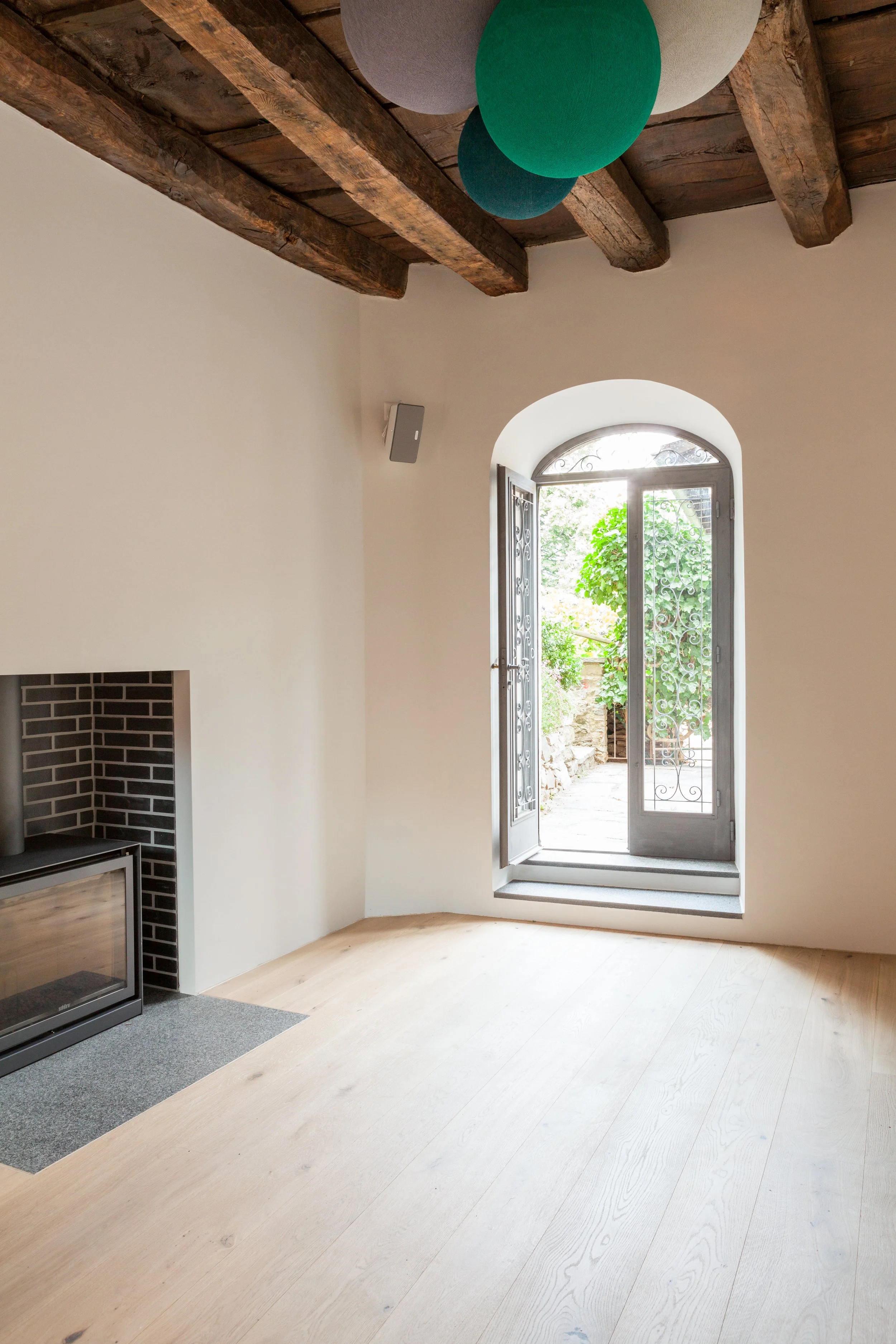 Stanza vuota con pareti bianche, pavimento in legno naturale, soffitto con travi in legno scuro e decorative sfere di carta colorate appese, finestra con inferriata e apertura su giardino verde