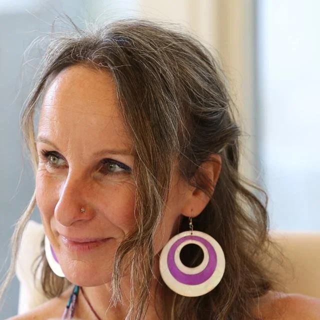 Donna con orecchini grandi bianchi con cerchi viola e neri, capelli ondulati, vicino a una finestra luminosa.