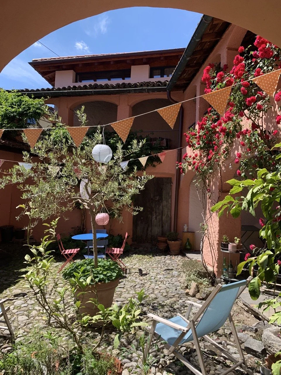 Interno di un cortile decorato con bandiere arancioni e lanterne colorate, piante, un tavolo blu con sedie rosse, una sedia a sdraio blu e vari fiori.