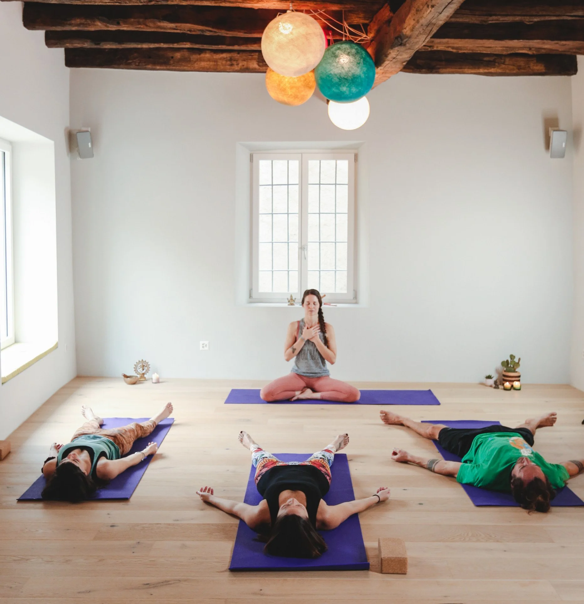 Una classe di yoga con quattro persone in posa supina e una donna in posizione di meditazione sullo sfondo, in una stanza luminosa con soffitto in legno e grandi finestre.