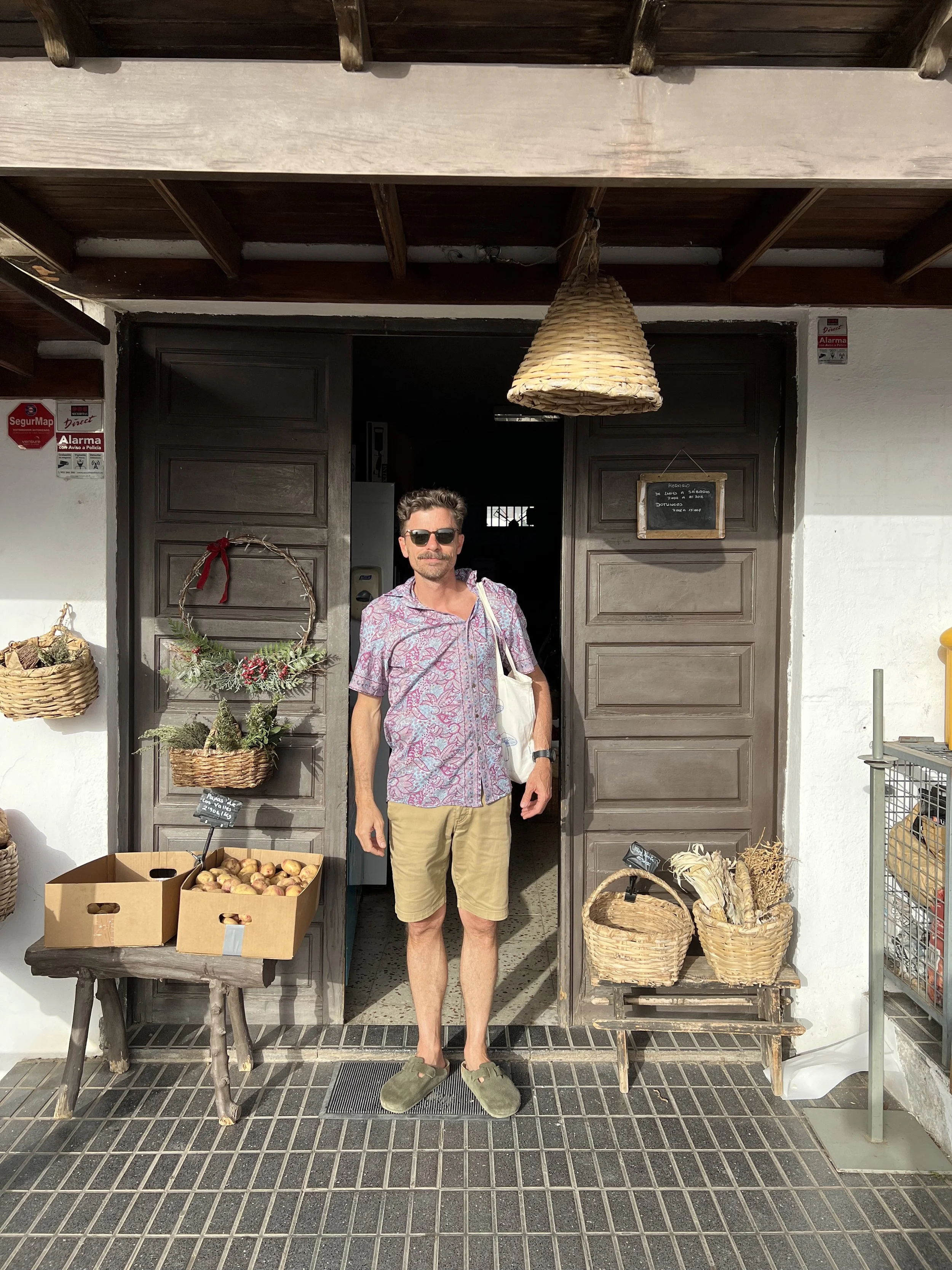 Uomo con occhiali da sole, maglietta a fiori, pantaloncini beige e slippers verdi, che soggiorna davanti a un negozio con decorazioni di natale e diviso da vasi con piante e con scatole di cipolle