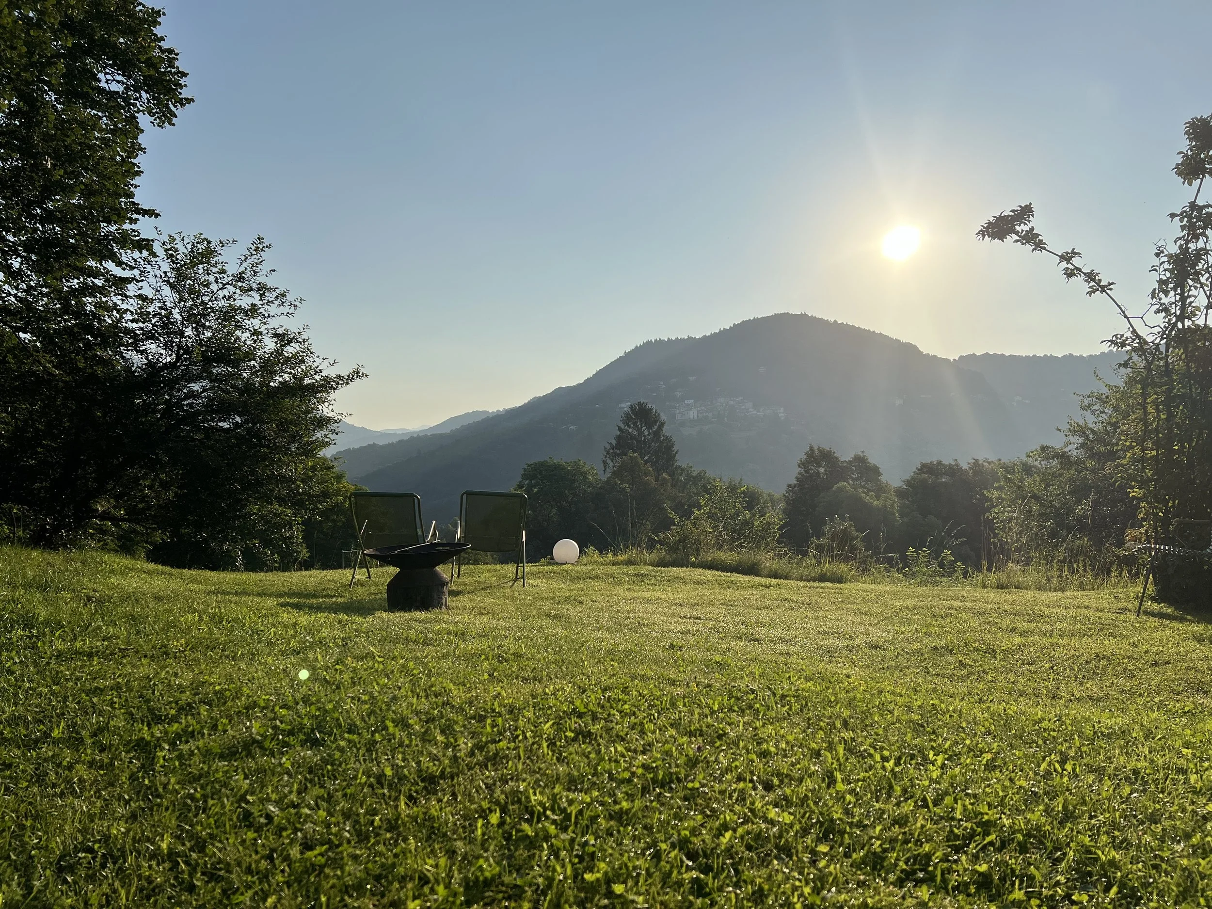 Campo verde con sedie, tavolino e lampada in primo piano, montagne sullo sfondo, sole nel cielo e alberi ai lati.