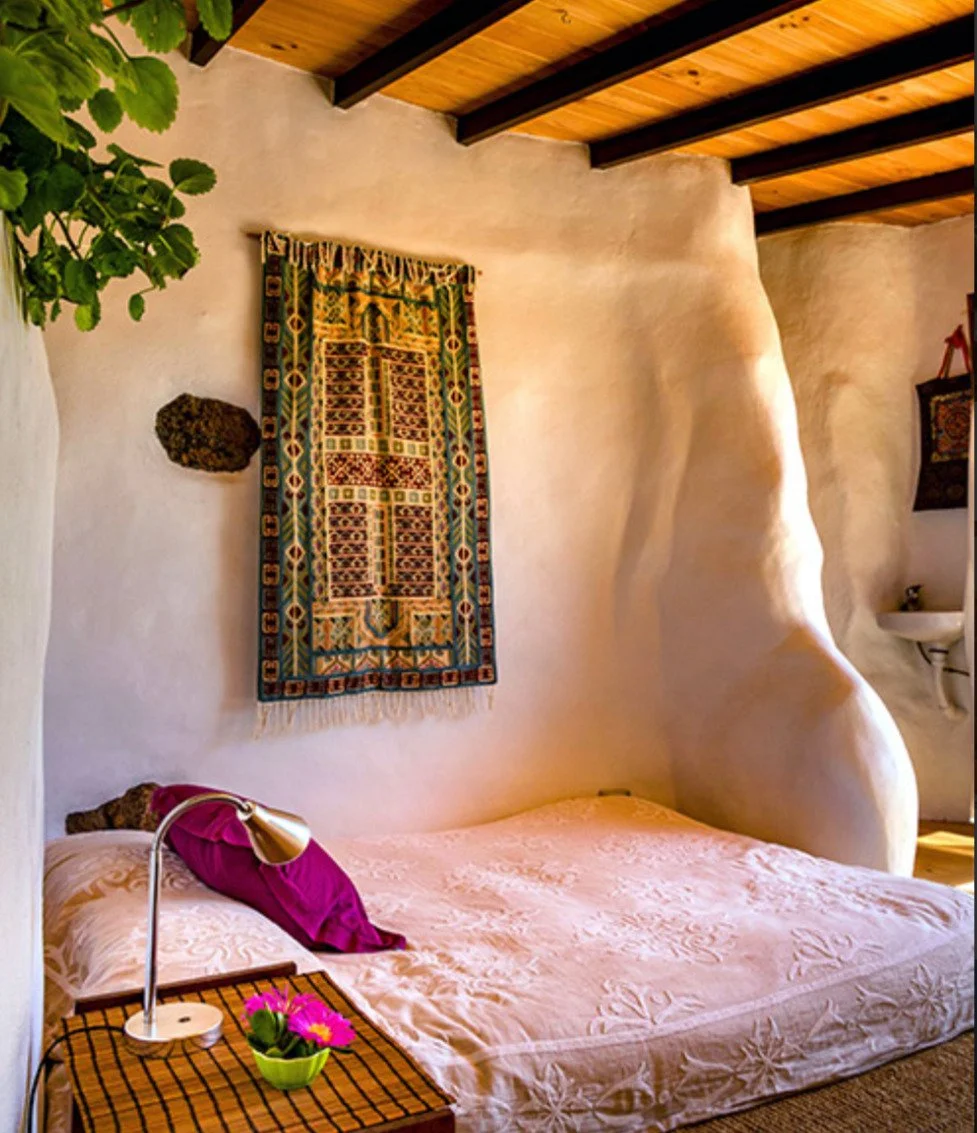Camera da letto con pareti in calce, decorata con un arazzo colorato, letto con coperta rosa e cuscino fucsia, lampada da tavolo, pianta in vaso e alcuni oggetti decorativi.